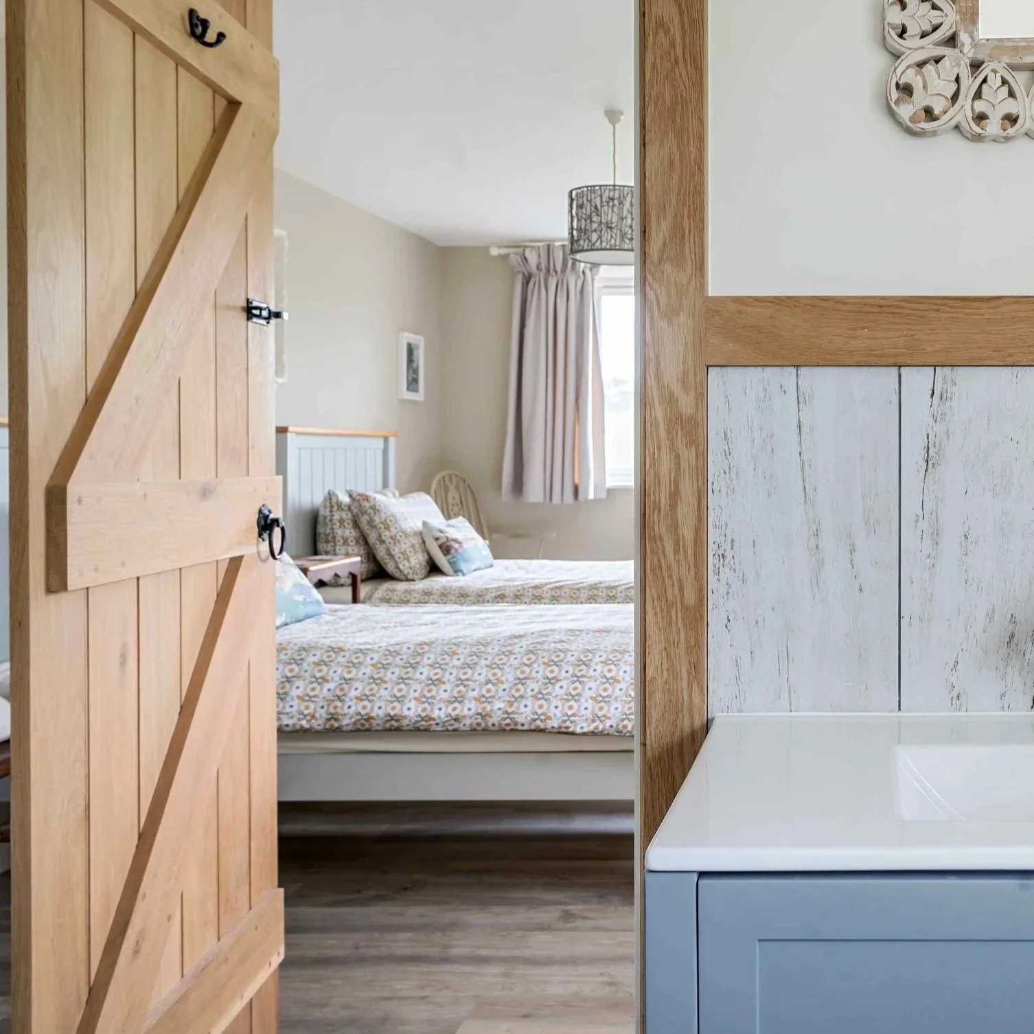 Interior of a room with a bed and a wooden sliding door on the left; a bathroom sink with a faucet in the foreground; a mirror and wall socket on the right wall with light-colored walls, wood trim, and patterned bedding and curtains.