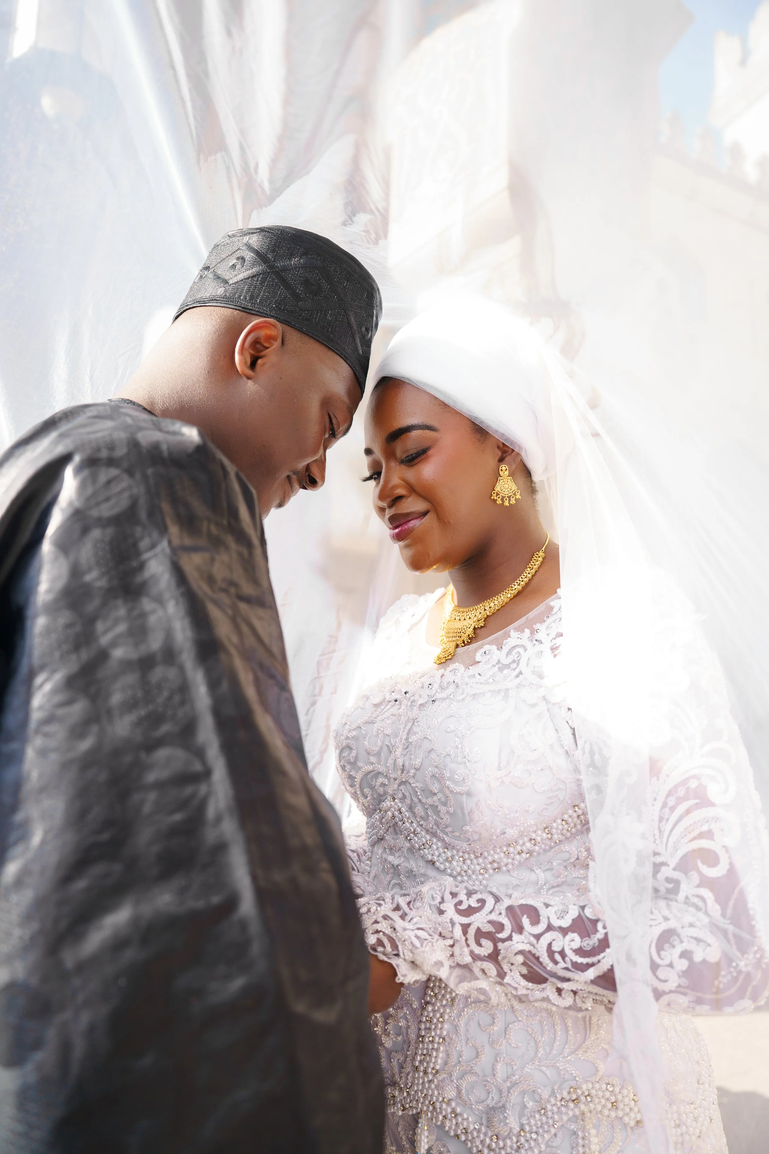 A man and woman in traditional attire sharing an intimate, tender moment on their wedding day. The woman is dressed in a white lace wedding dress with intricate embroidery and gold jewelry, while the man wears a dark traditional outfit and a cap. They are leaning close, smiling gently at each other in a brightly lit setting.