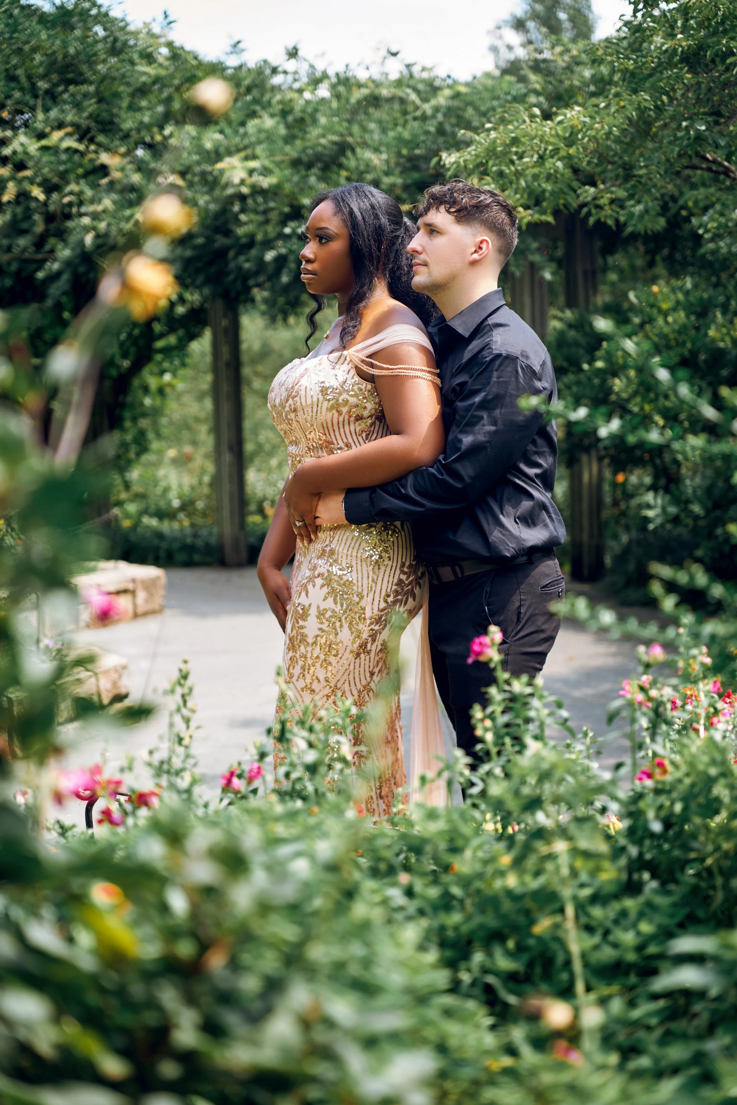 A bride dressed in white in a lace wedding gown and headscarf stands under a stone archway, with a man dressed in traditional black Nigerian attire nearby, in an outdoor setting with trees and flags in the background.