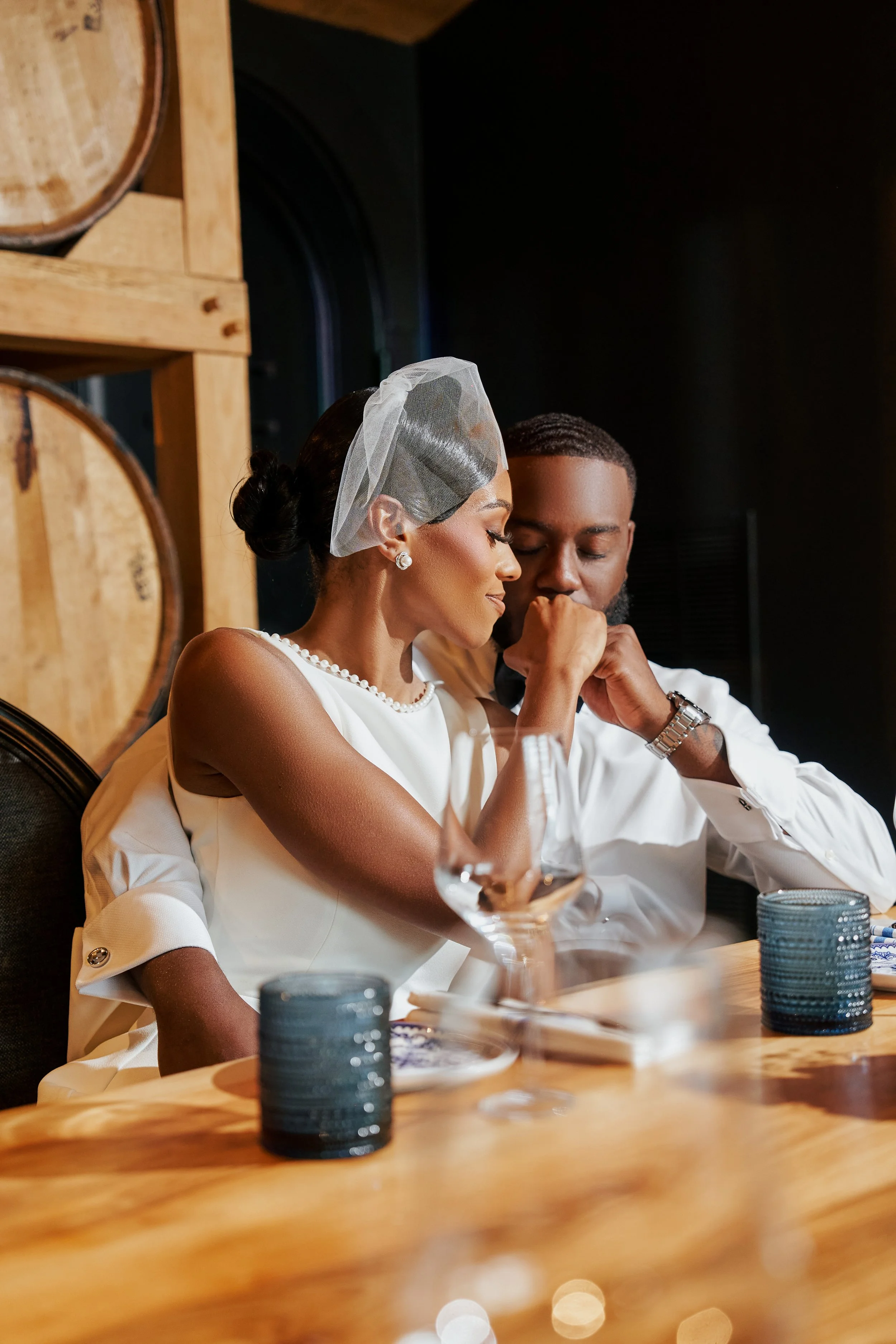 A woman and a man are sitting at a wooden table, holding hands and touching foreheads in an intimate moment. The woman is dressed in a white dress with pearl details and is wearing a hair net. The man is wearing a white shirt and a watch.