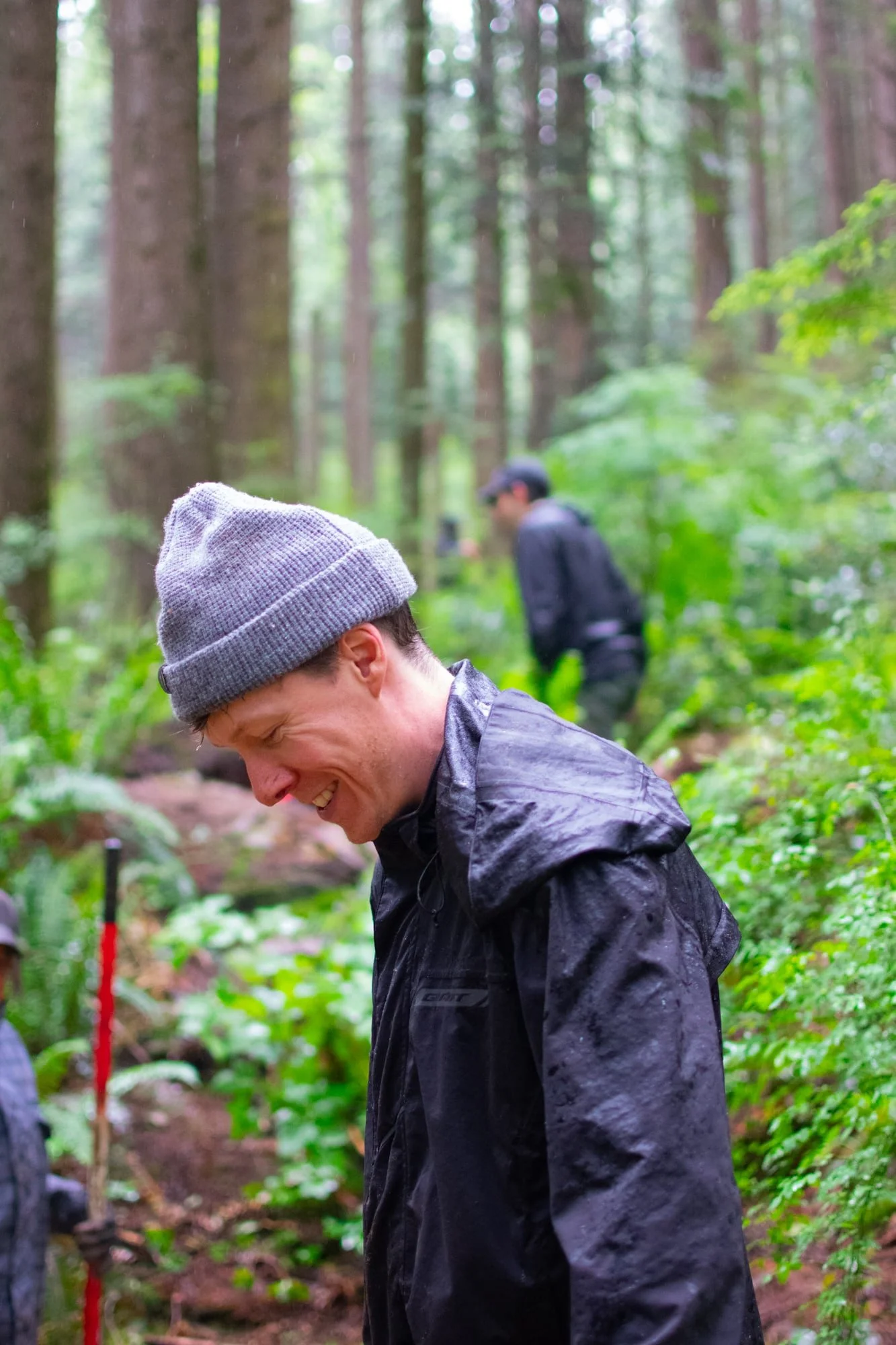 Person in a gray beanie and raincoat in a forest setting, smiling, with another person blurred in the background.