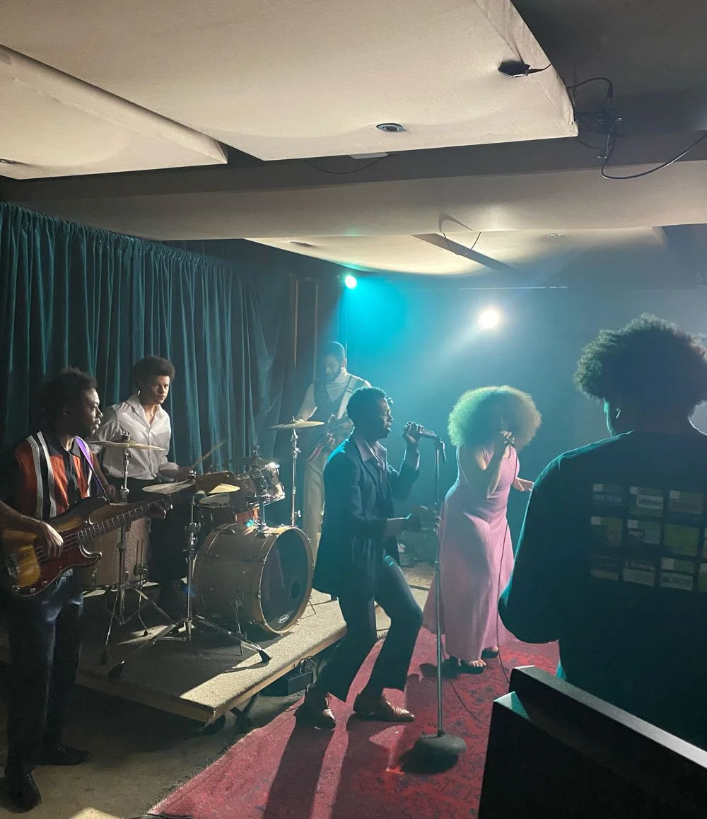 A live music performance featuring a band on stage, with a singer holding a microphone and other musicians playing instruments, illuminated by colorful stage lights, in a club or small venue setting.