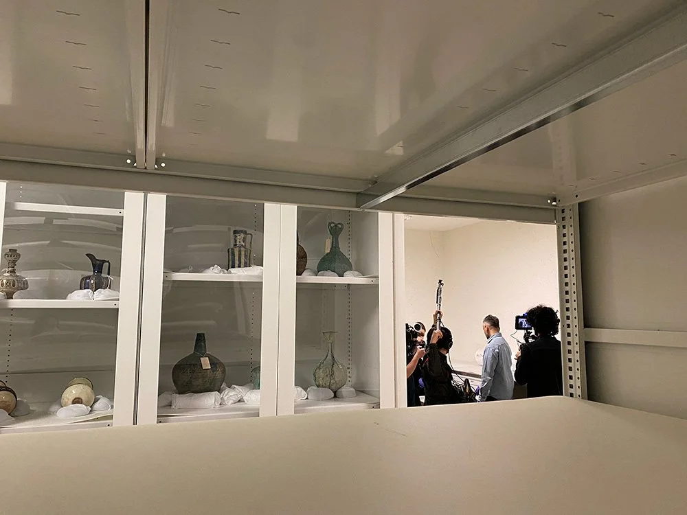 A group of people, including camera operators, filming or photographing a man in a blue shirt in a room with white shelves containing various ceramic vases.