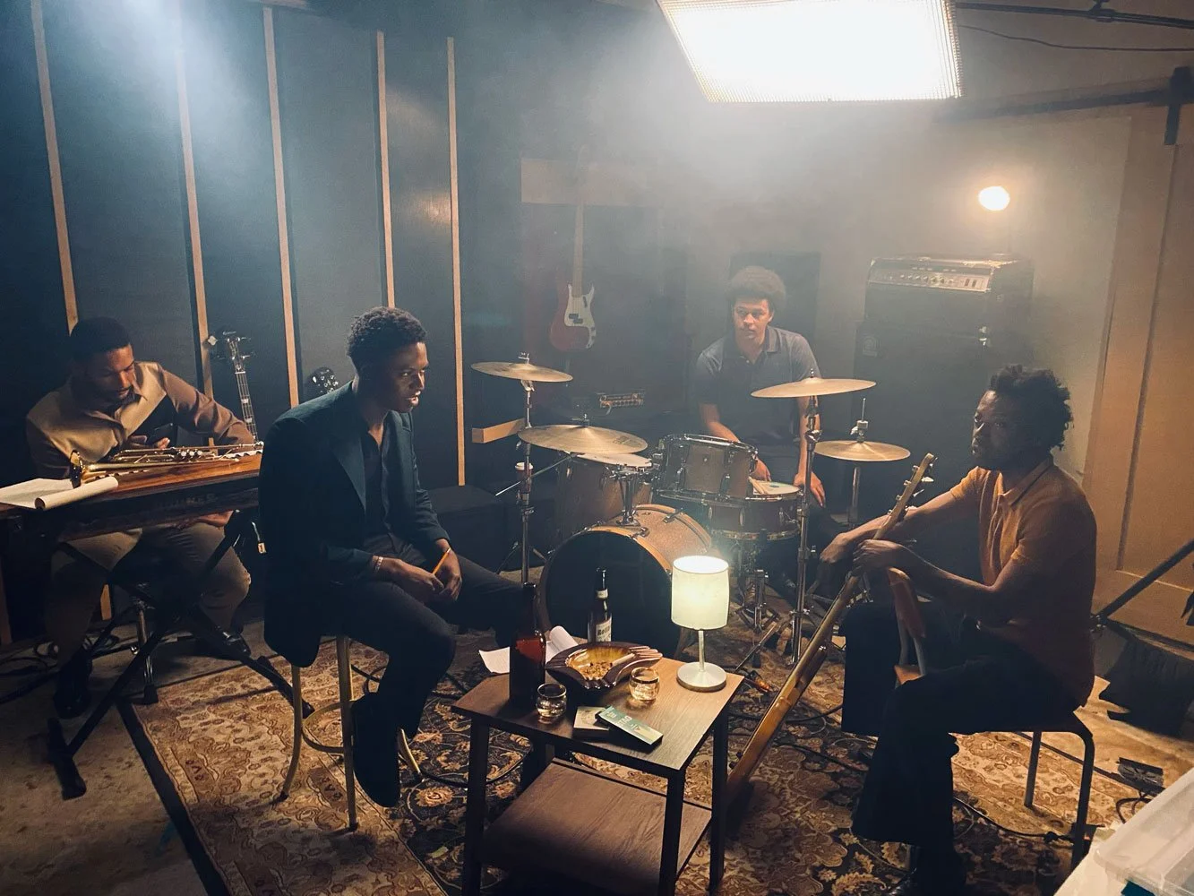 Four musicians practicing in a studio with a drum set, and guitar, illuminated by a small lamp with food and drinks on a table.