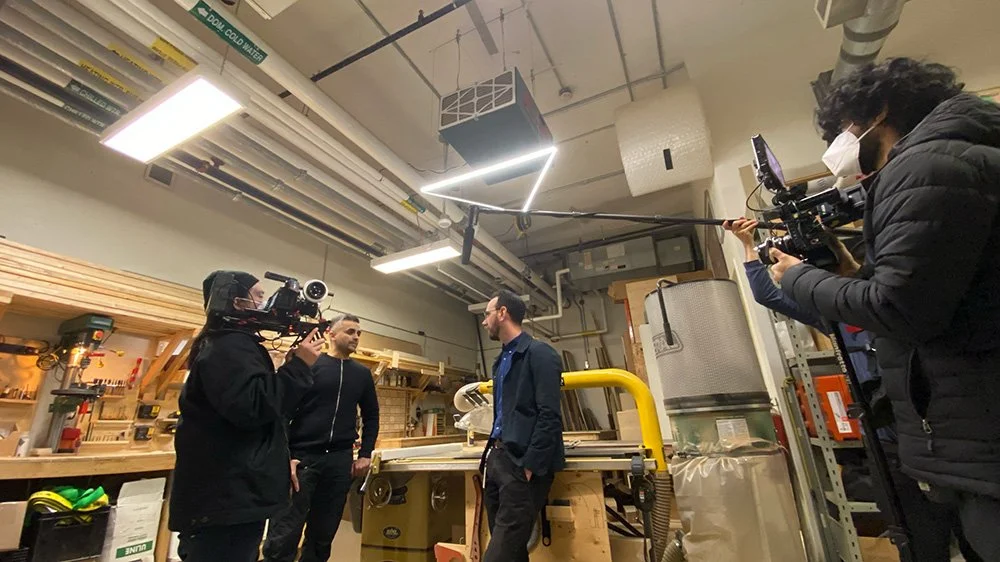 Two camera operators filming a man in a blue jacket and dark pants in a woodworking shop or workshop, with tools and equipment in the background.