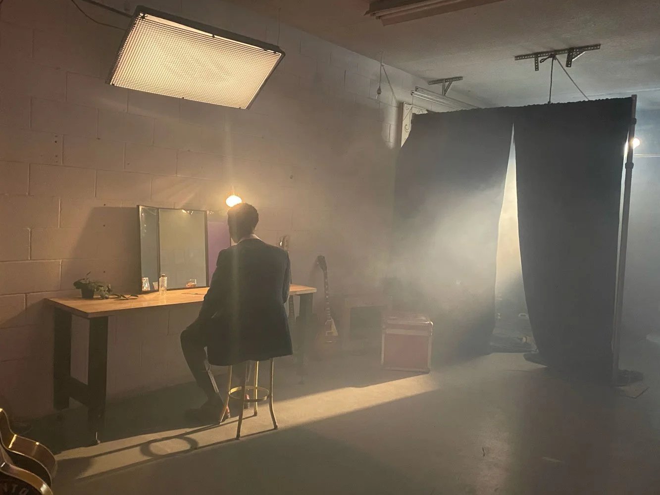 A man in a suit sitting on a stool in front of a mirror, in a dimly lit room with a fabric backdrop, some equipment, and a guitar in the background, with sunlight filtering through the curtain.