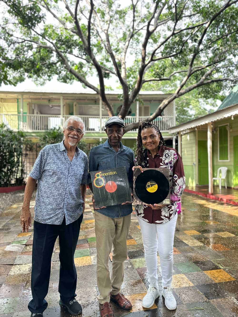 Three people stand outside on a patio holding vinyl records. A large tree with green leaves is in the background along with a building with a balcony and another building with green siding. 