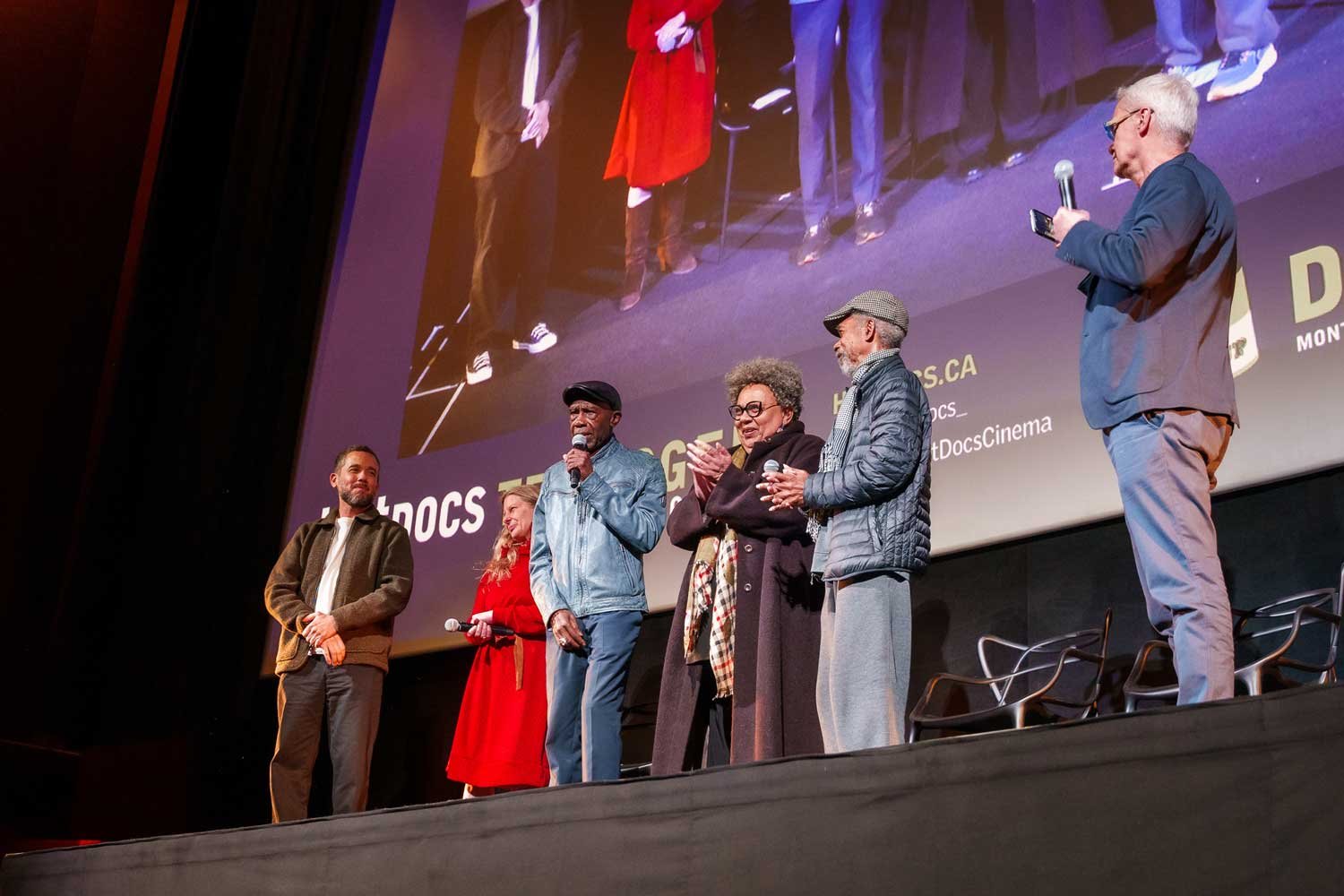 Six people standing on a stage in front of a large screen during an event, some holding microphones, one person looking at their phone, the background shows their reflection on the screen.