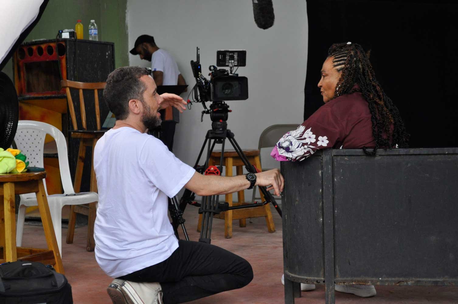 A Director kneeling and talking to a woman sitting on a chair on a film set with a camera on a tripod filming them.