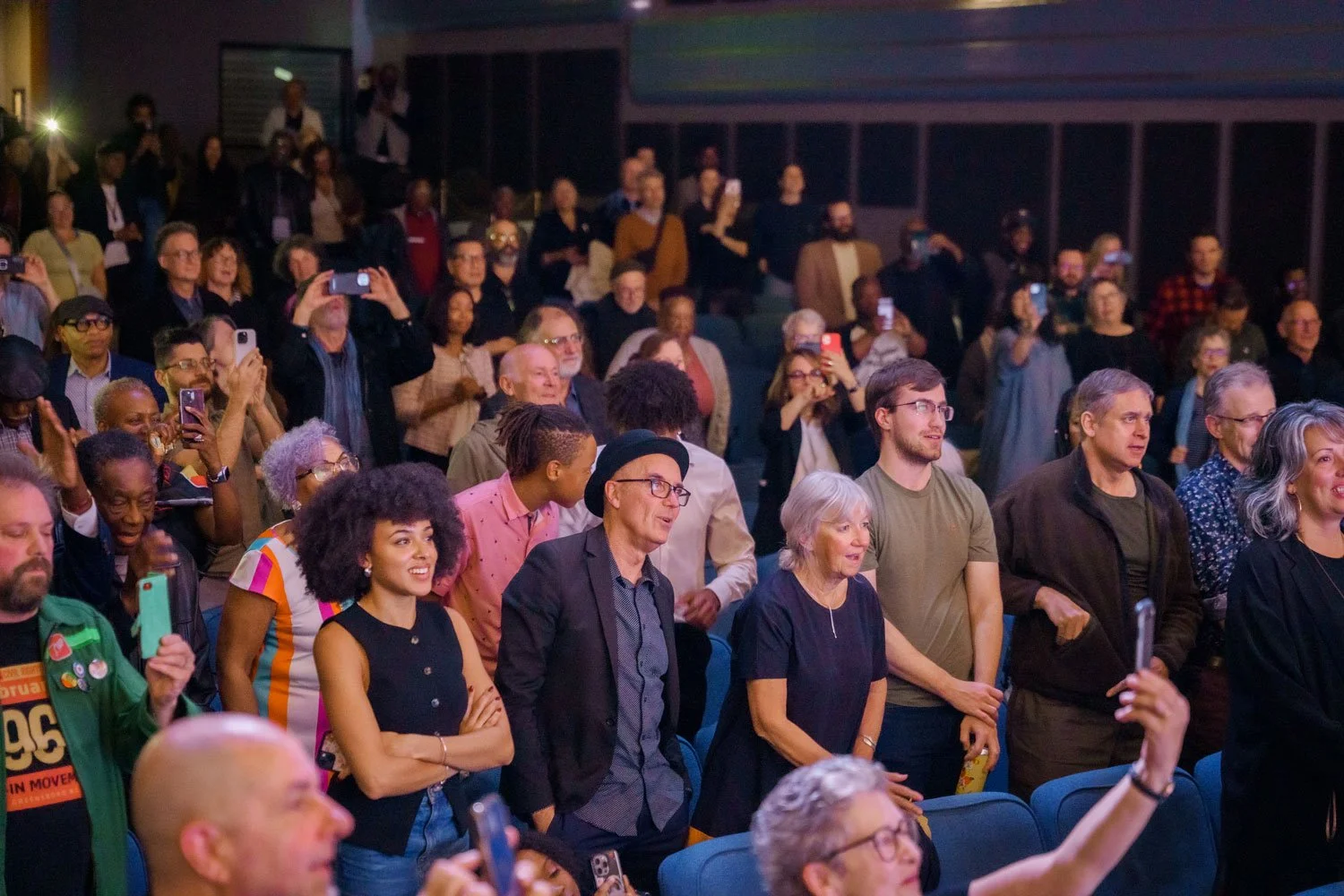 A diverse group of people gathered at an indoor event, many of them standing and taking photos or videos with their phones, with some seated and engaged, in a dimly lit environment.