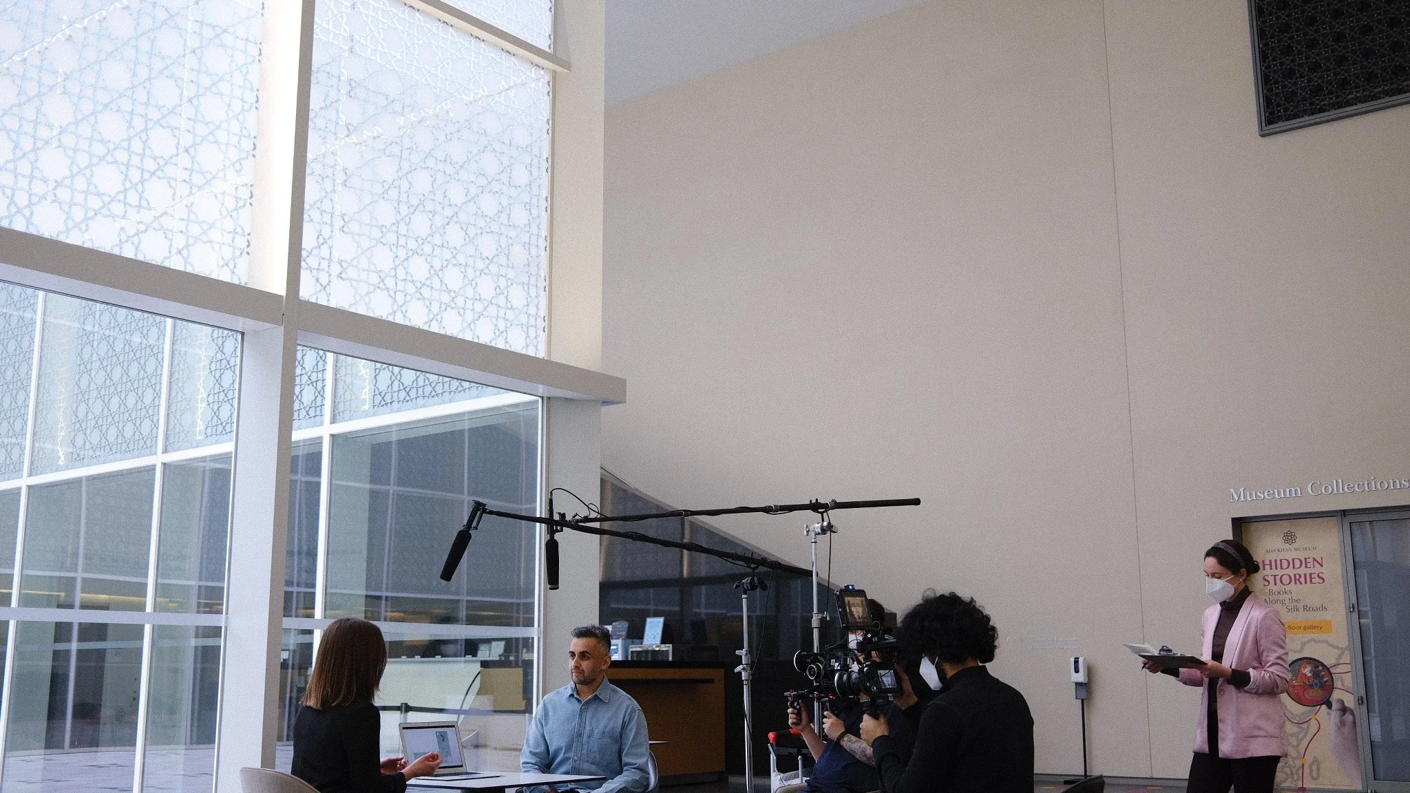 Inside a modern museum or gallery with large windows and geometric patterned glass, people are engaged in a discussion or interview with recording equipment present, and a woman wearing a face mask is reading from a paper.