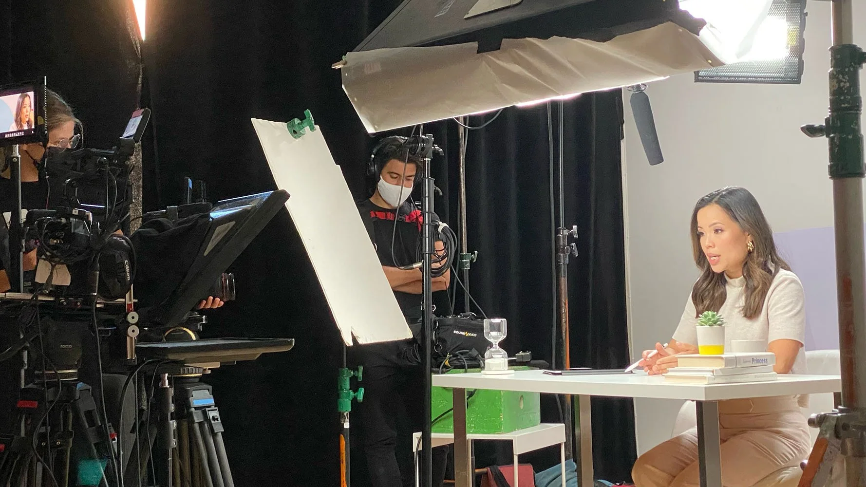 A woman on set of a video shoot filmed indoors, sitting at a table with books and a potted plant, while a crew member wearing a mask appears to be adjusting camera equipment.