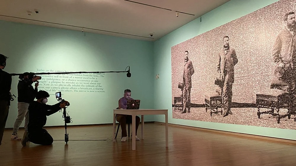 A person working at a desk with a laptop in an art gallery, filming a man sitting at the desk, with a large pixelated artwork of four men in suits on the wall behind.