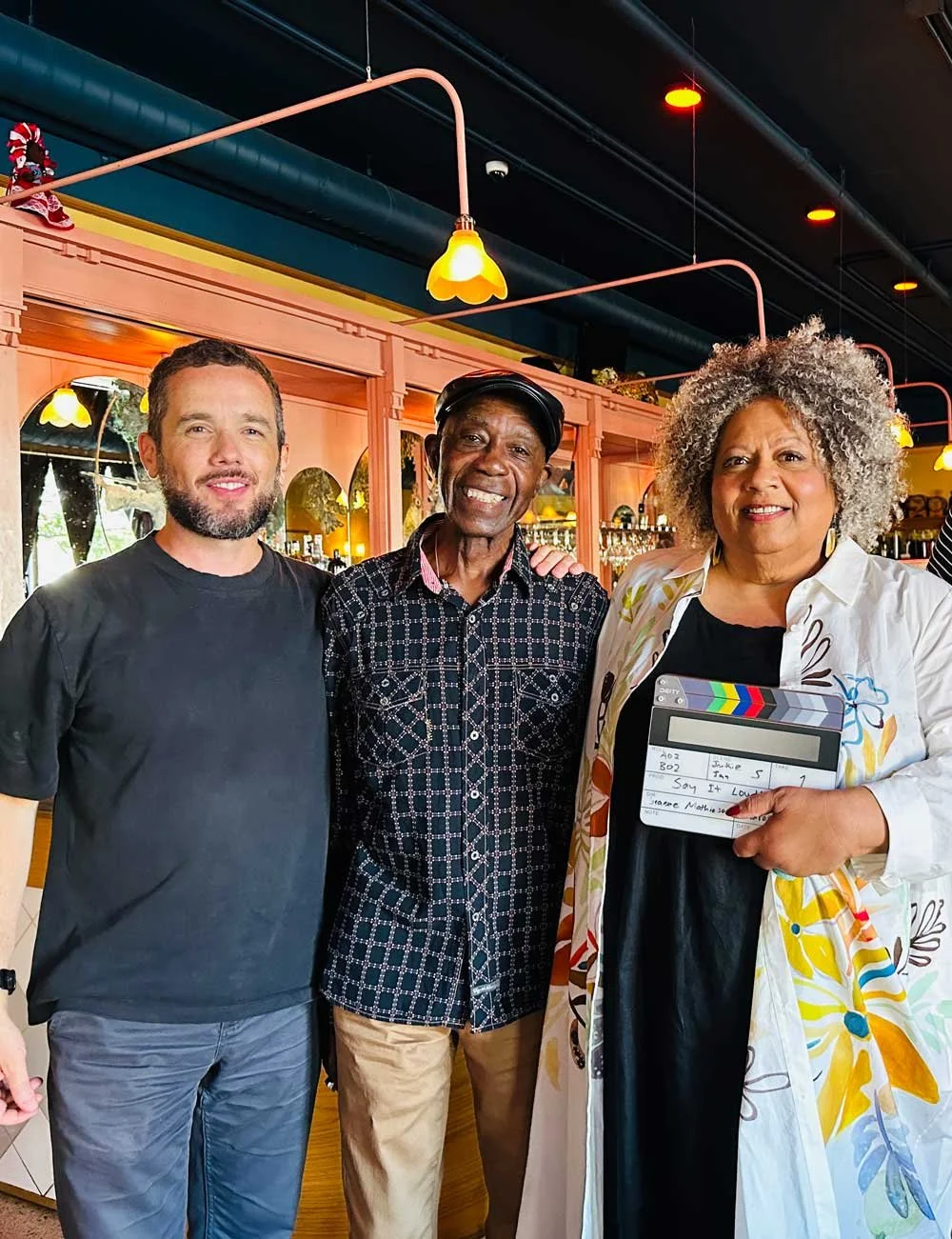 Three people standing together inside a restaurant, smiling. The person on the right holds a clapperboard, and the background shows warm lighting and bar shelves.