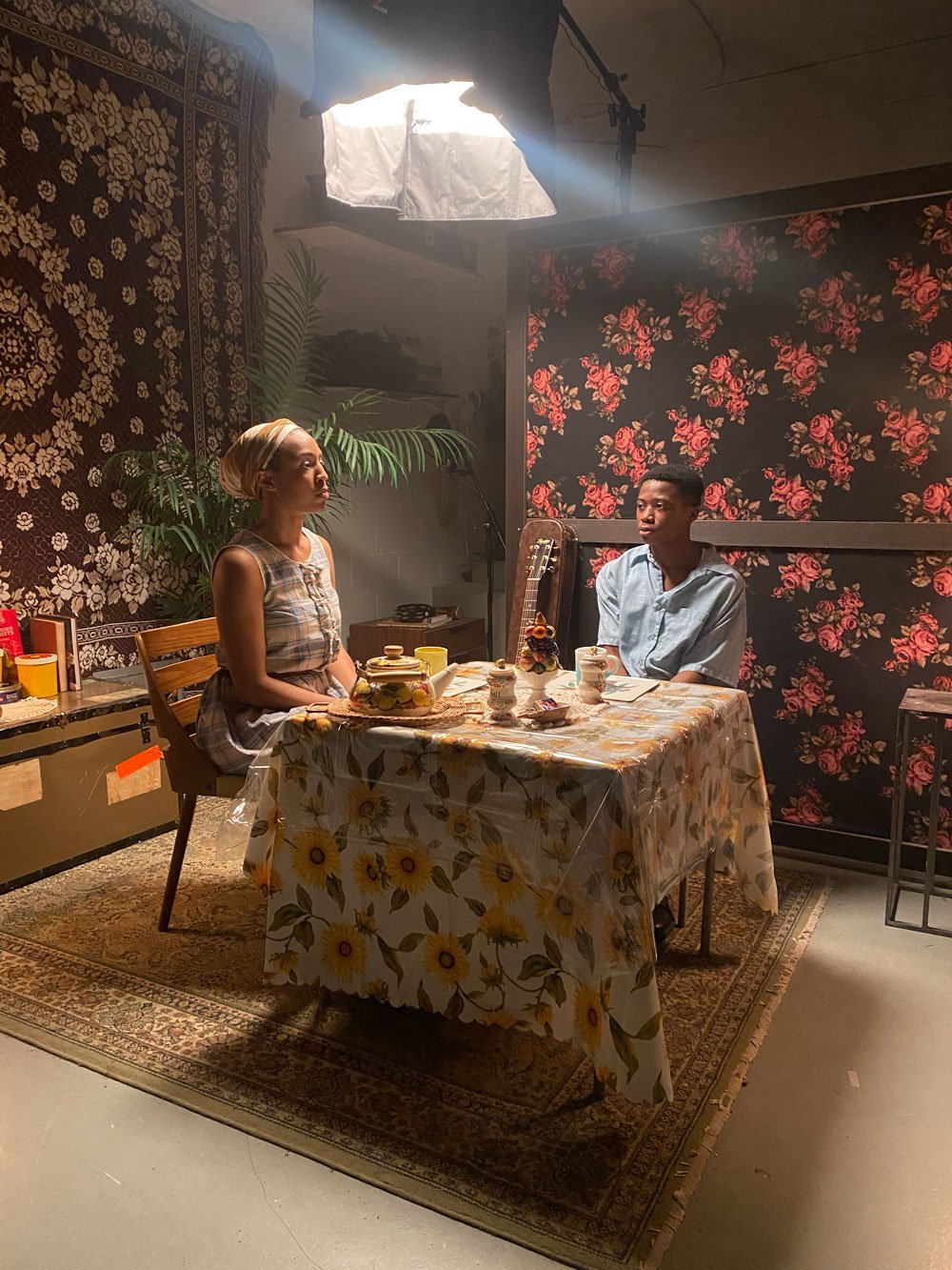 Two people sitting at a dining table in a room with floral patterned wallpaper. One woman in a sleeveless dress and headwrap on the left, and a man in a light blue shirt on the right. There is studio lighting equipment above them, and various cups an