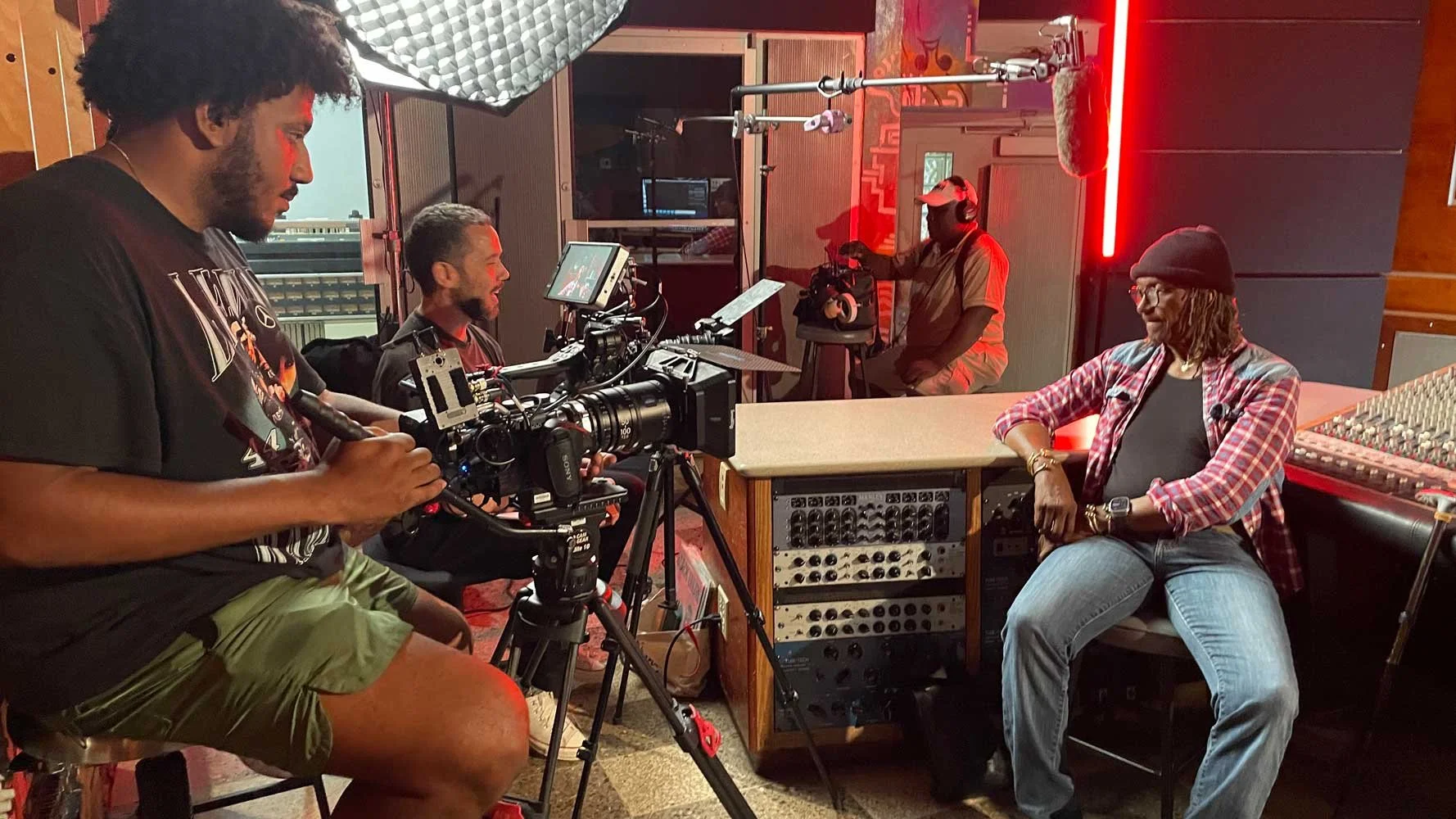 A behind-the-scenes look at a recording session in a music studio. Four people are involved: one man is holding a camera on a tripod, filming a woman sitting at a mixing console. The woman is wearing a plaid shirt, black top, and gray jeans, and is s