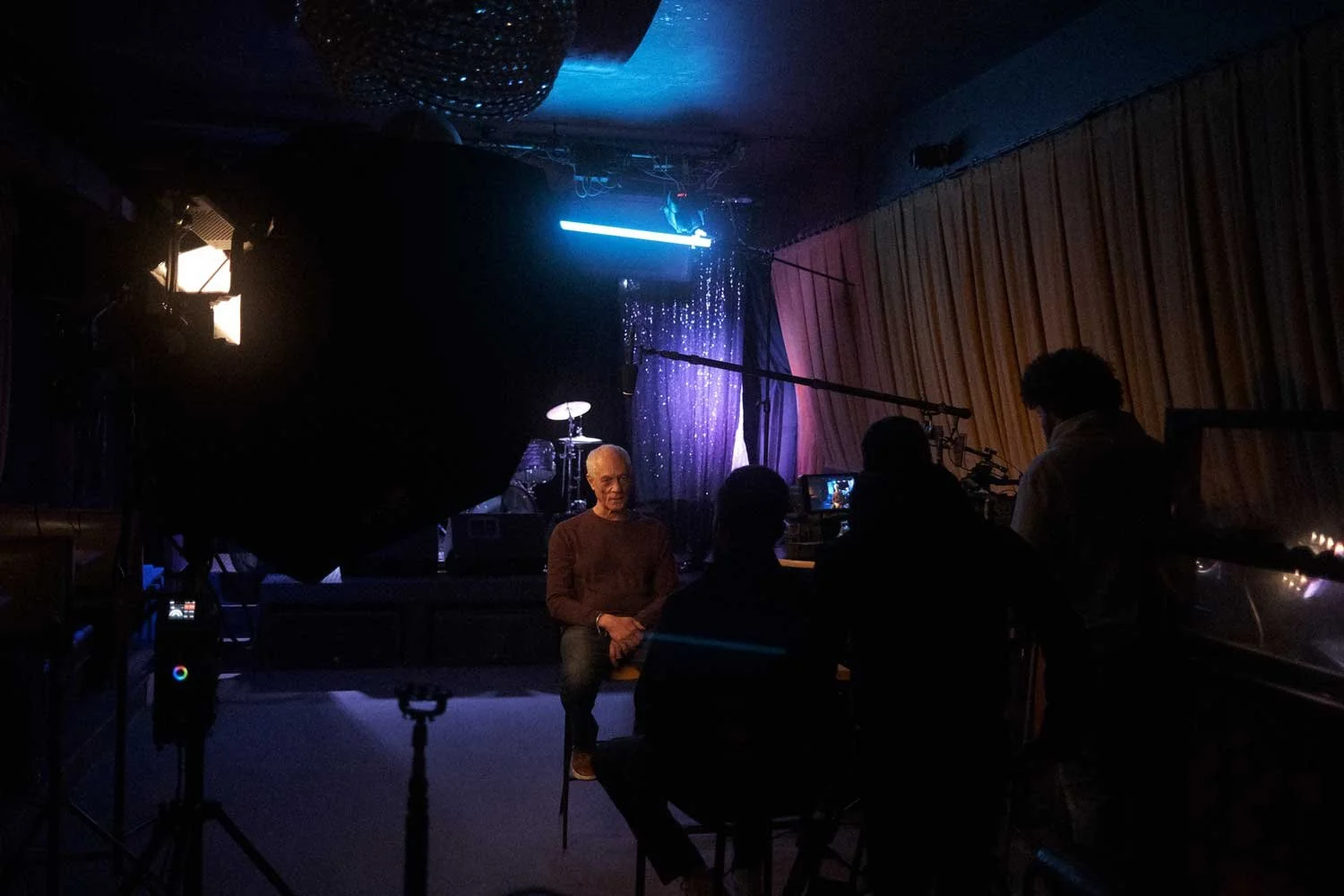An interview recording taking place in a dark room with a man sitting on a stool, with crew members operating camera equipment around him, and a drum set visible in the background.