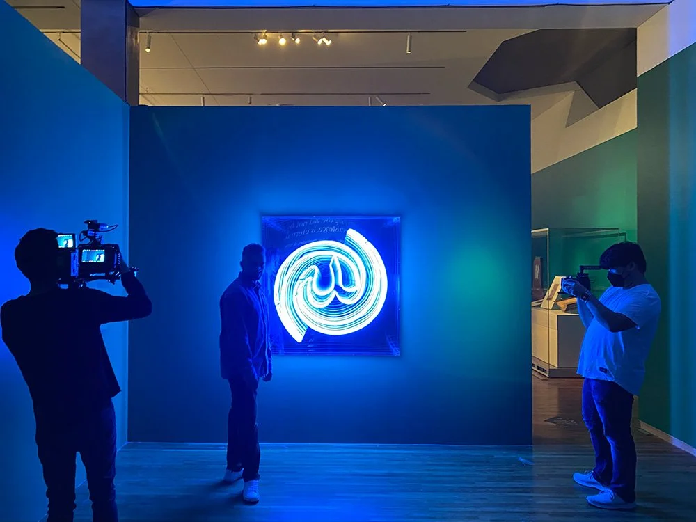 People taking photographs of a neon light artwork featuring a spiral design with the Twitter logo, displayed on a dark wall in an art gallery.