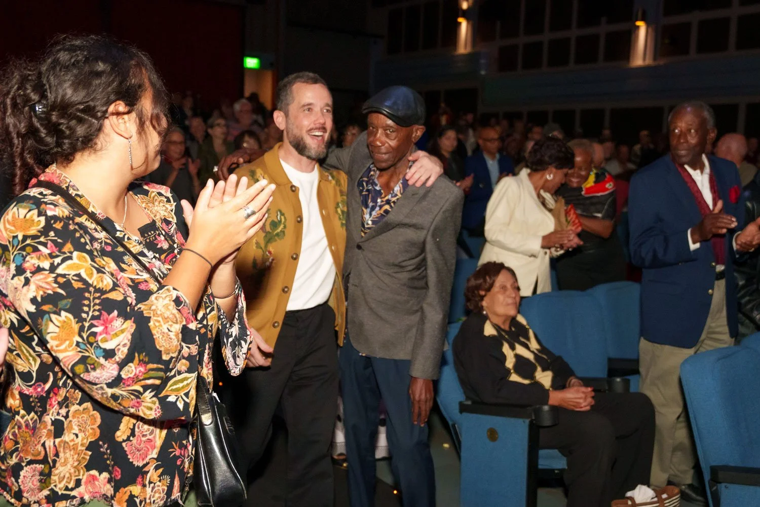 Photo from a documentary film premiere. People at an event, with two men in the center smiling and embracing, while others around them are standing, clapping, and looking on.