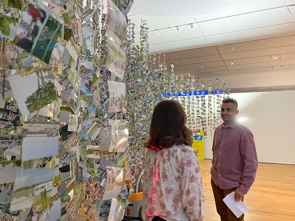 Two people, a man and a woman, stand in front of a wall decorated with numerous photographs hanging from strings in an art gallery. The man is smiling, facing the woman, who has her back to the camera. The gallery has wood flooring and a white backgr