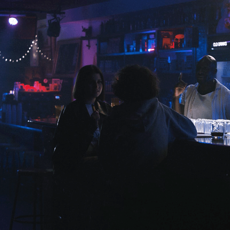 Three people in a dimly lit bar, two sitting at the counter and one bartender standing behind it, with shelves of liquor bottles in the background.