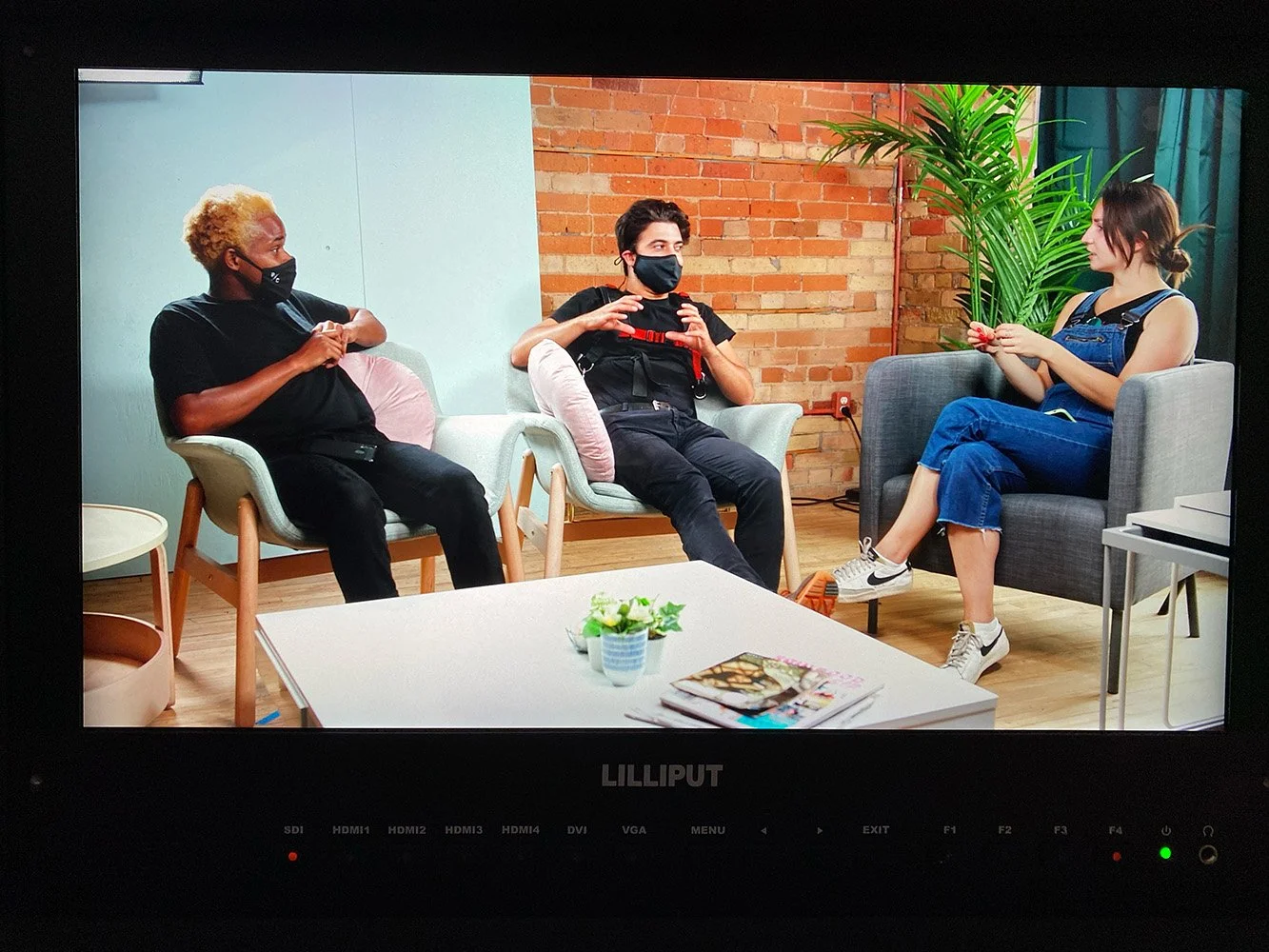Three people sitting and talking in a studio with a brick wall and a large plant in the background. Two women and one man, all wearing face masks, seated on chairs around a coffee table with magazines and a small potted plant.