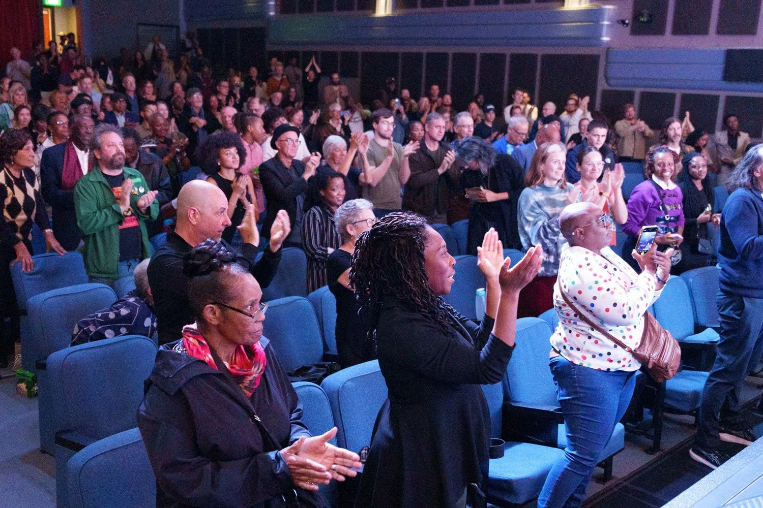 Crowd of diverse people standing and clapping in an indoor venue, some taking photos or videos.
