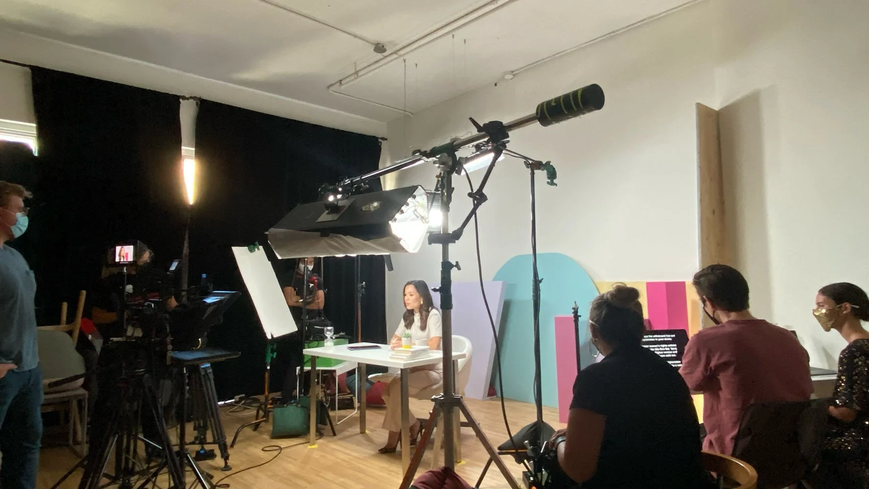 Indoor filming studio with a woman seated at a table, cameras and lighting equipment, crew members, and several people seated watching a presentation. Black curtains cover the windows, and colorful background panels are behind the woman.