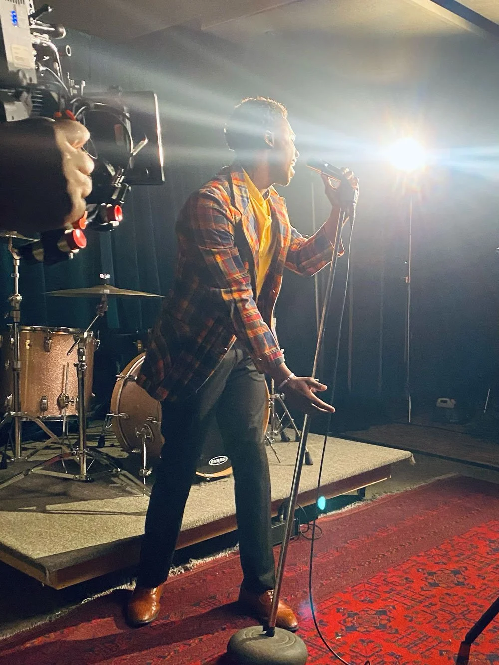 A man singing into a microphone on stage with a bright light shining behind him, a drum set in the background, and a camera filming him.