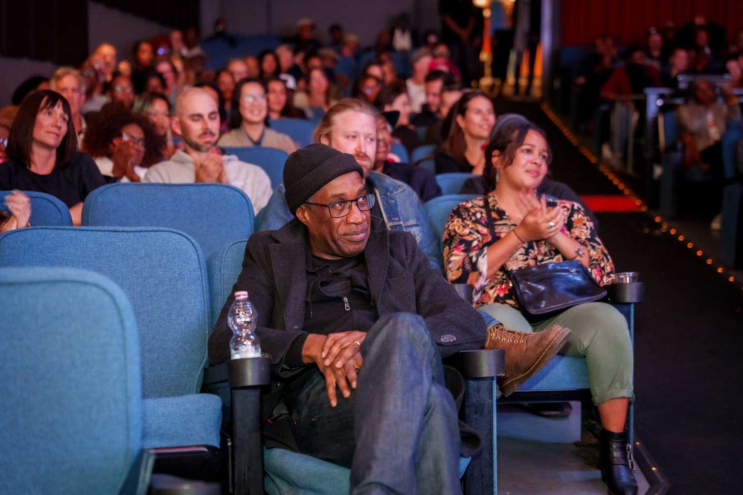 People sitting in theater seats attending a performance or event, with some clapping and watching attentively.