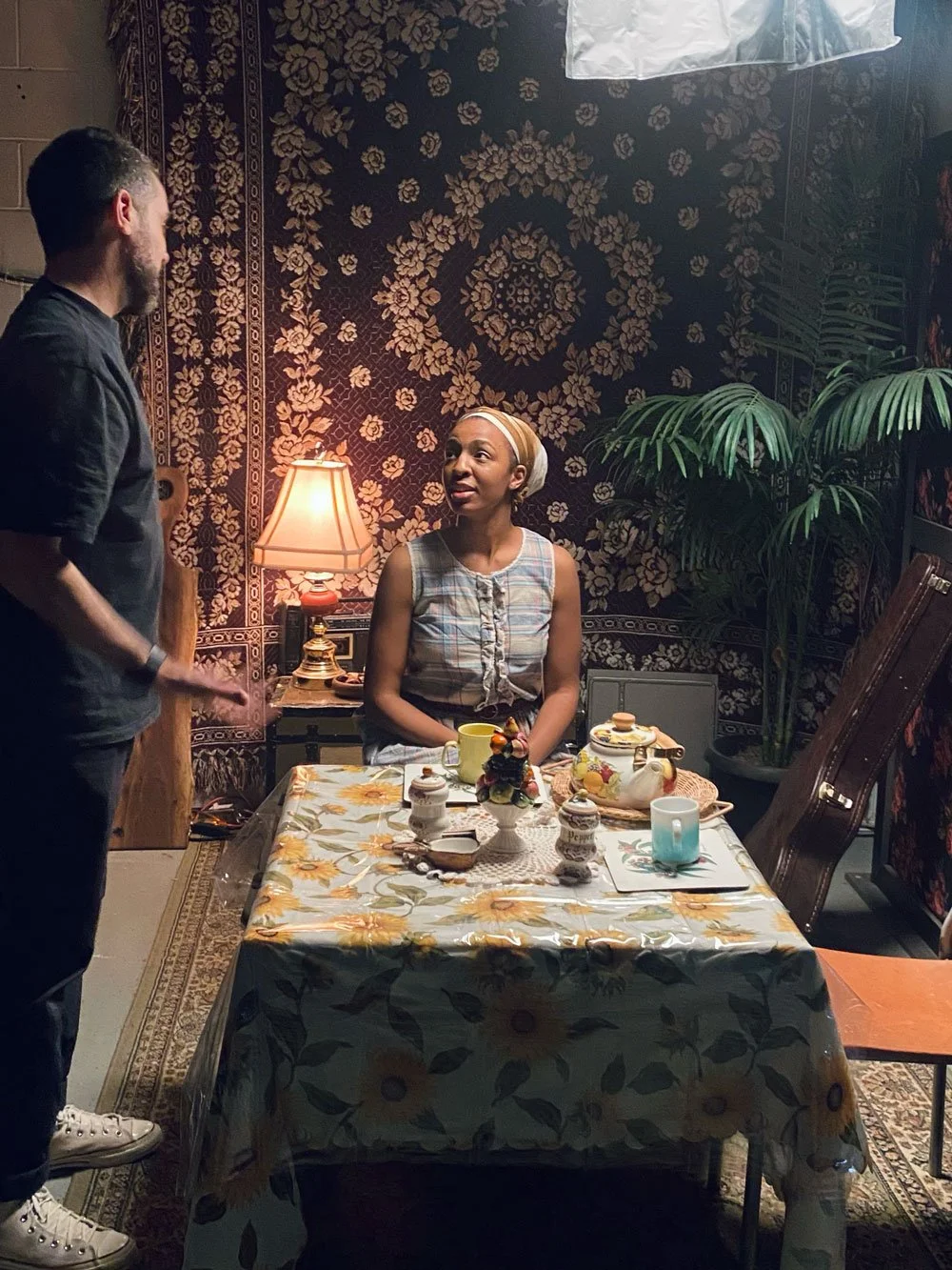 A woman sitting at a cluttered dining table with decorative items, cups, and a teapot as she looks up at a man standing nearby in a room with floral and patterned wallpaper, a table lamp, and a large green plant in the background.