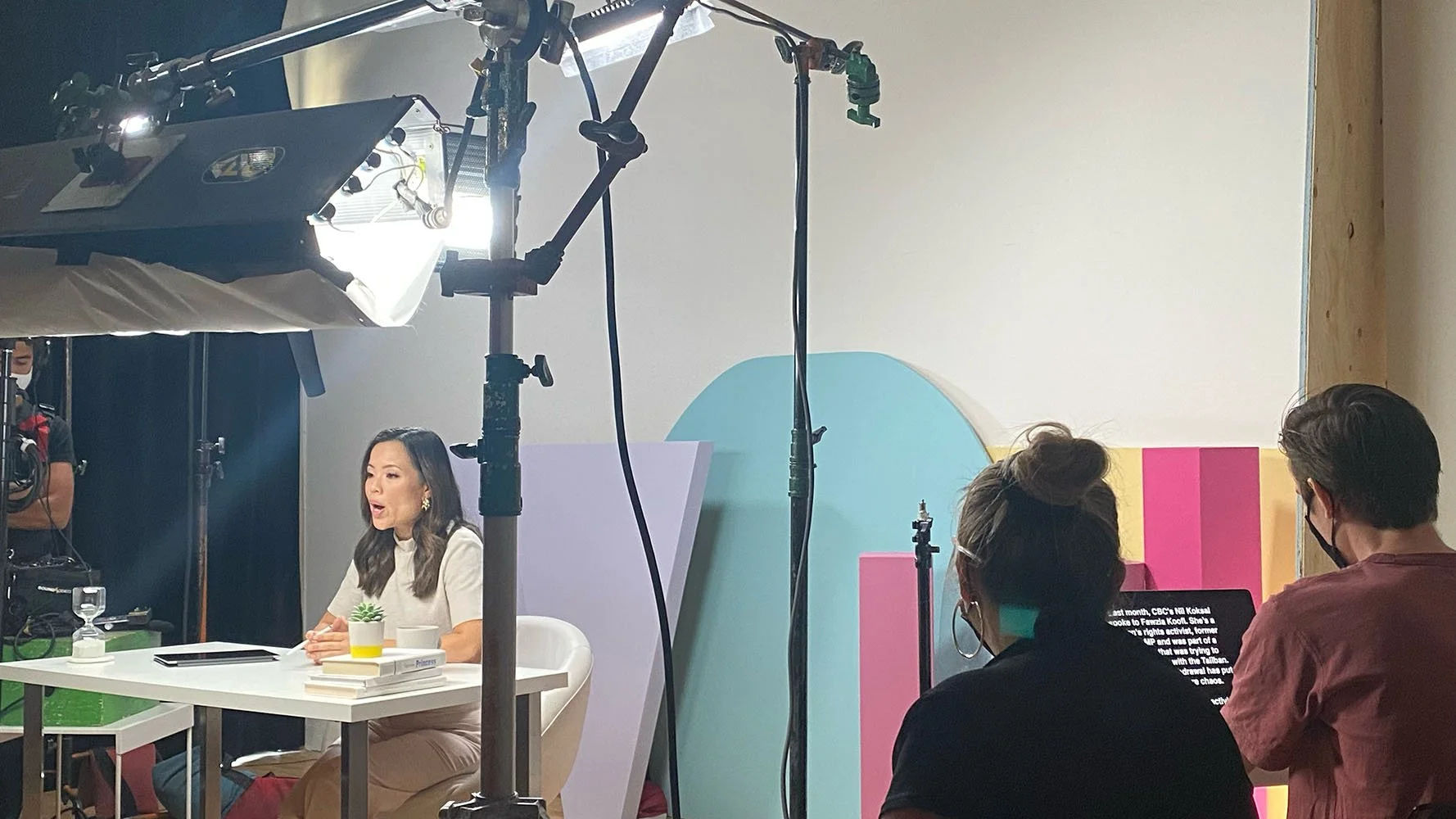 A woman is sitting at a table with a tablet, papers, a glass of water, and a small potted plant, appearing to be in a recording studio or TV studio. There are three people in the foreground, two men and one woman, watching her. Equipment and colorful