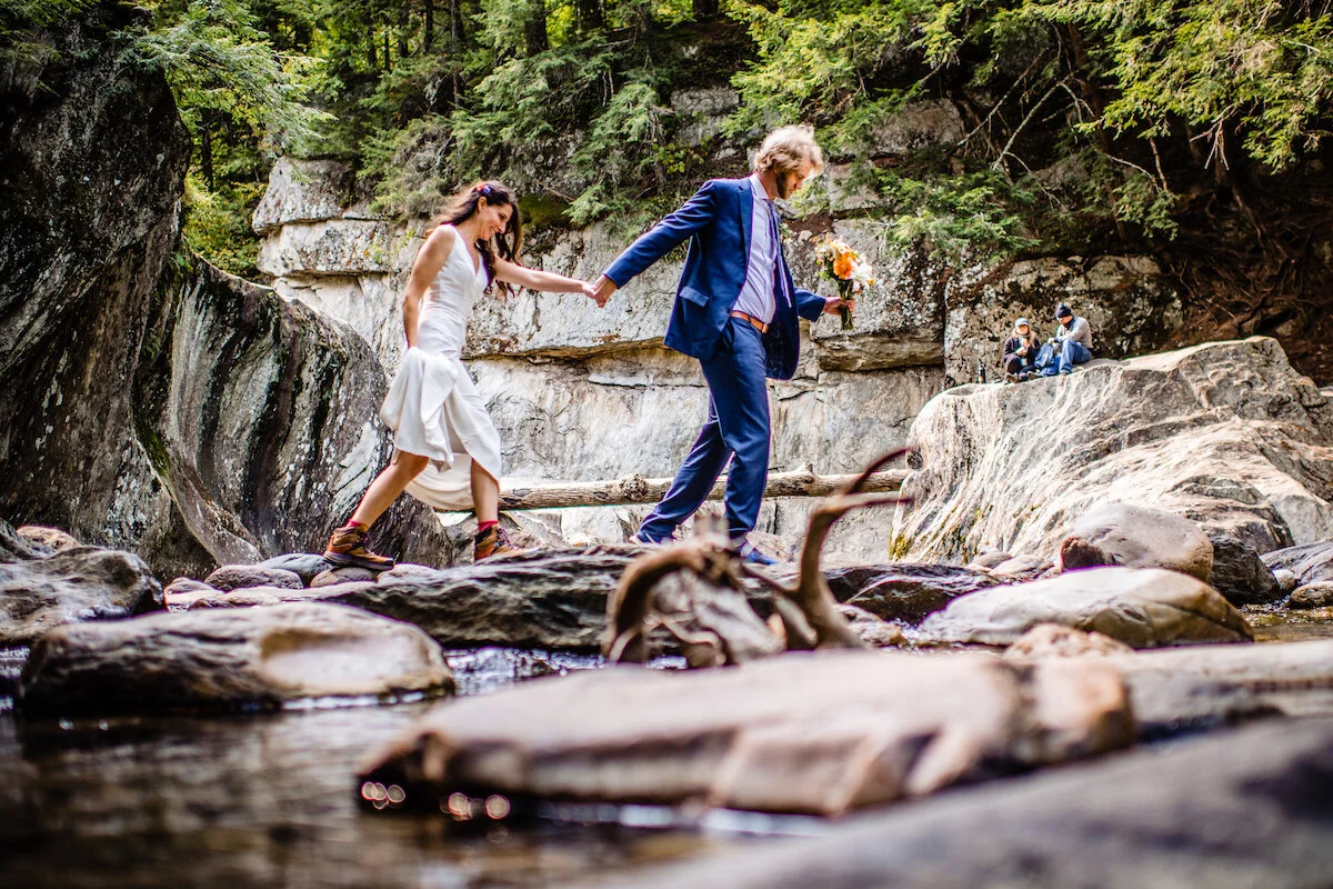 Groom leads bride over creek and mountain rocks