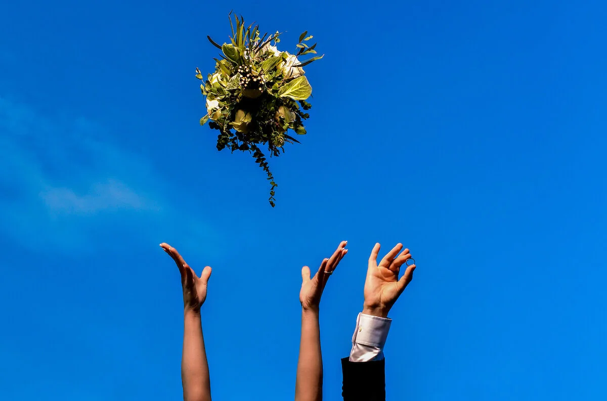 Bride throwing bouquet in while groom holds ring blue skies background