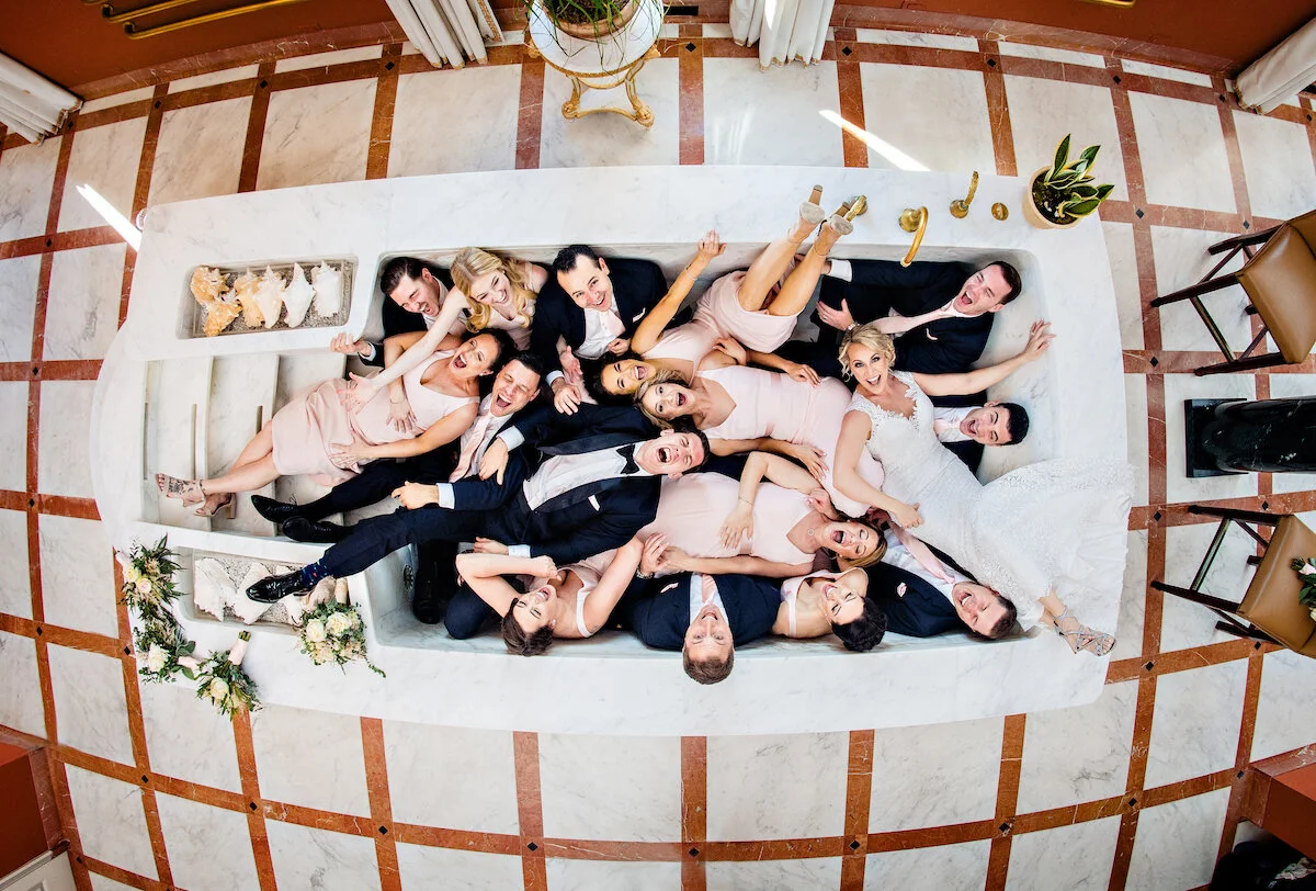 Groomsmen and bridesmaids with bride and groom posing in bridal suite bathtub