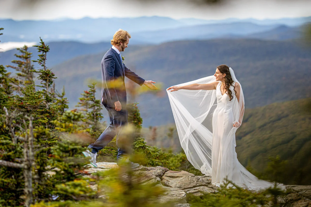 Bride and groom extend to hold hands with mountains in background