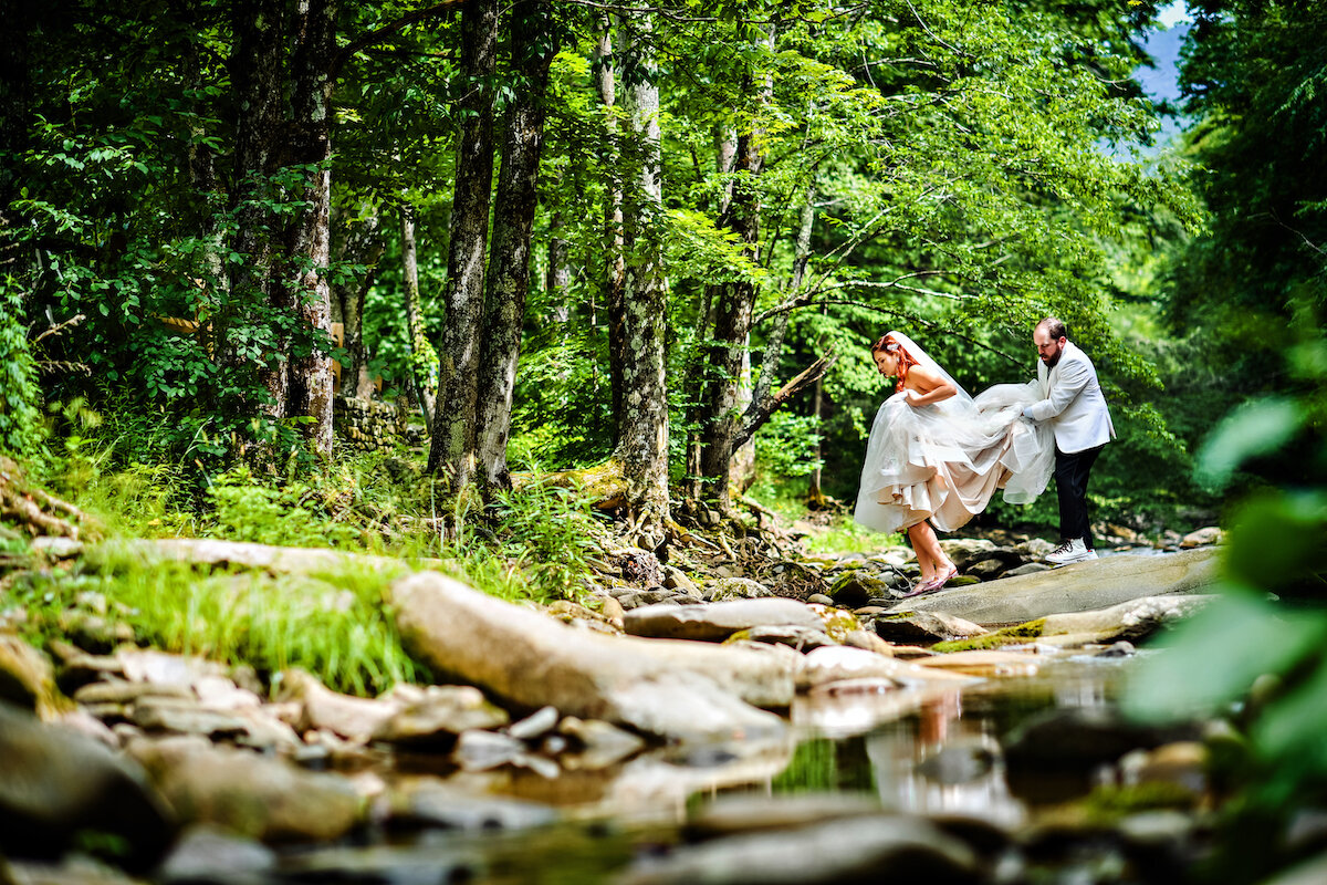 Groom holds brides dress as they walk over creek in forest