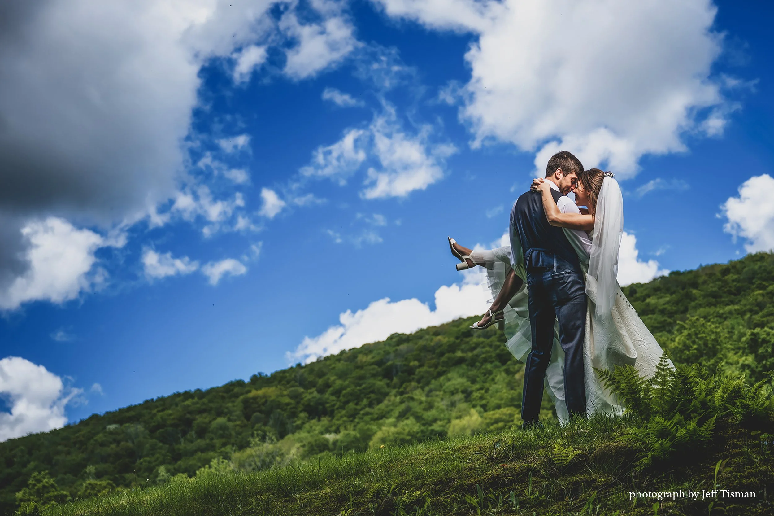 wedding couple embraces on a hillside under a bright blue sky with fluffy white clouds