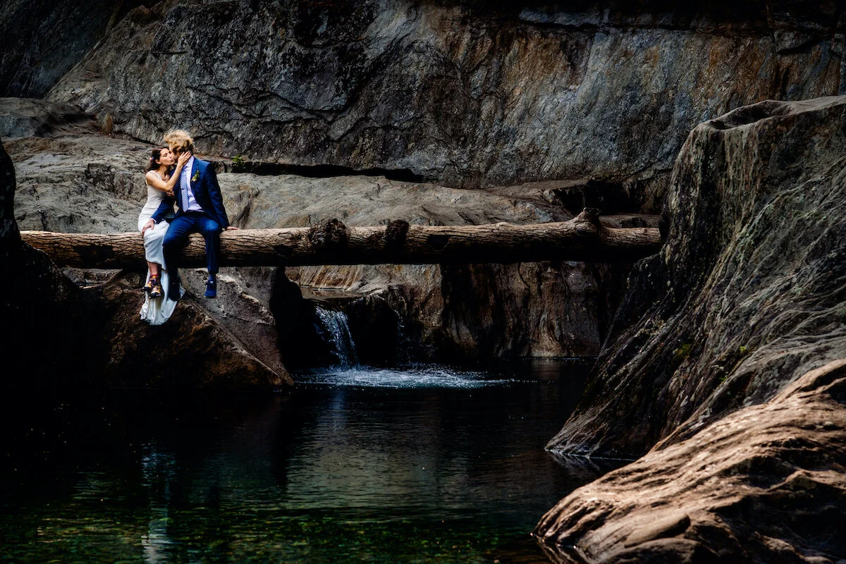 Bride and groom sit on fallen tree above mountain stream