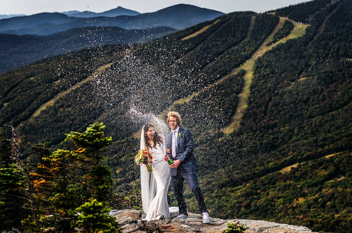 Bride and groom pop shaken champagne bottle at top of mountain