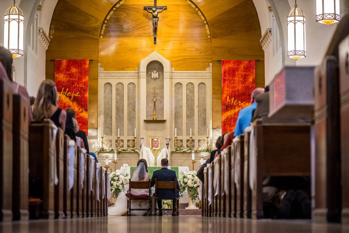 Bride and groom sit as priest blesses with view of guests in pews