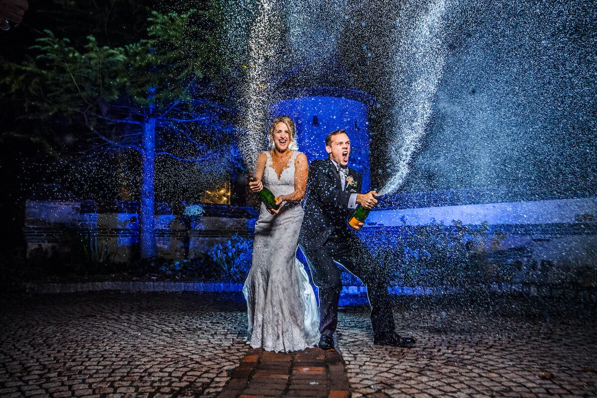 Bride and groom open shaken champagne bottles on cobblestone road