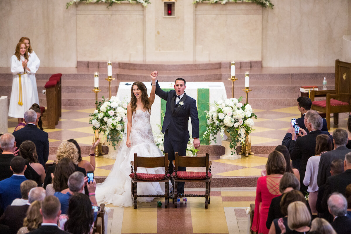 Bride and groom hold hands high as guests take pictures and cheer