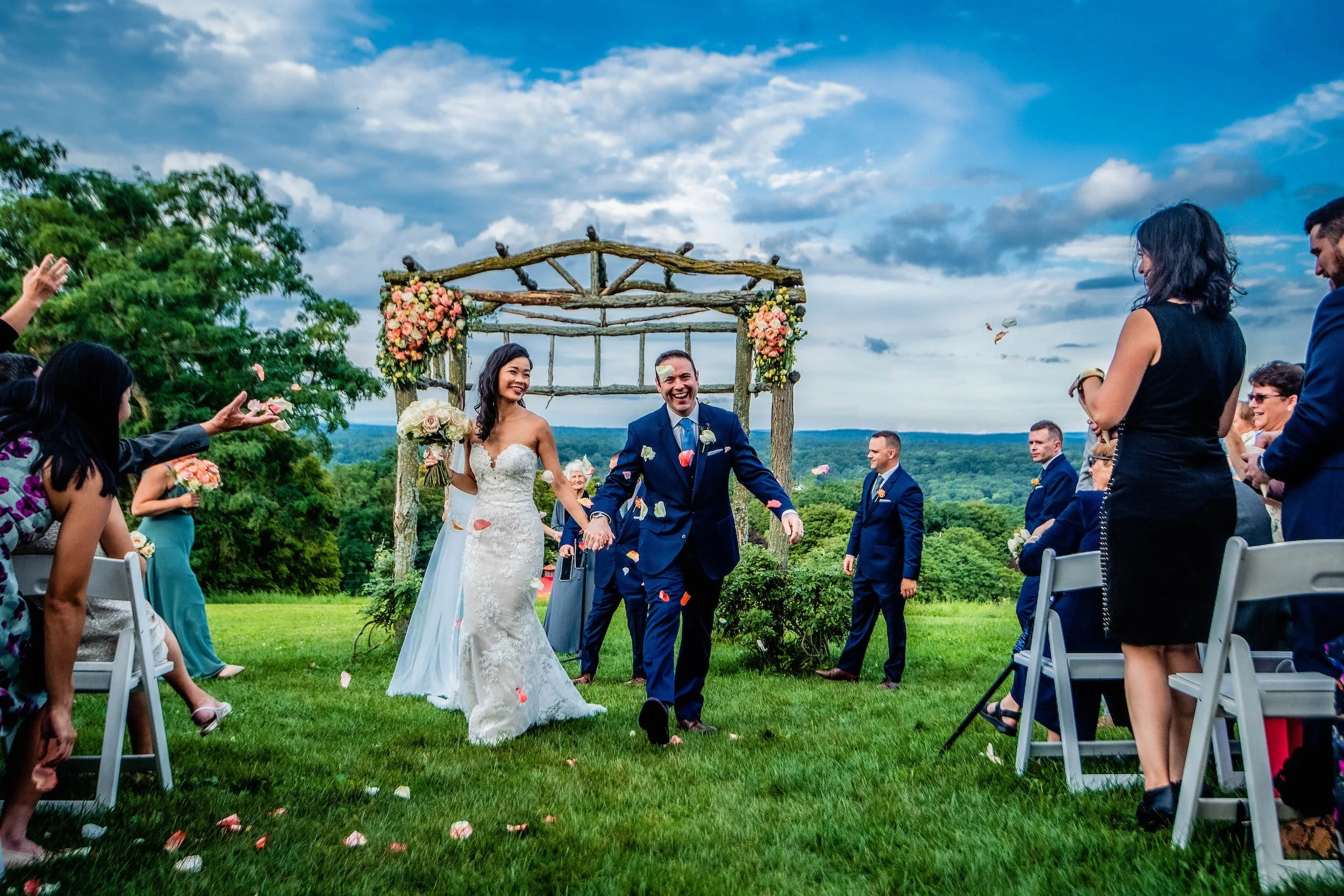 Bride and groom walk down aisle outdoors as wedding guests cheer