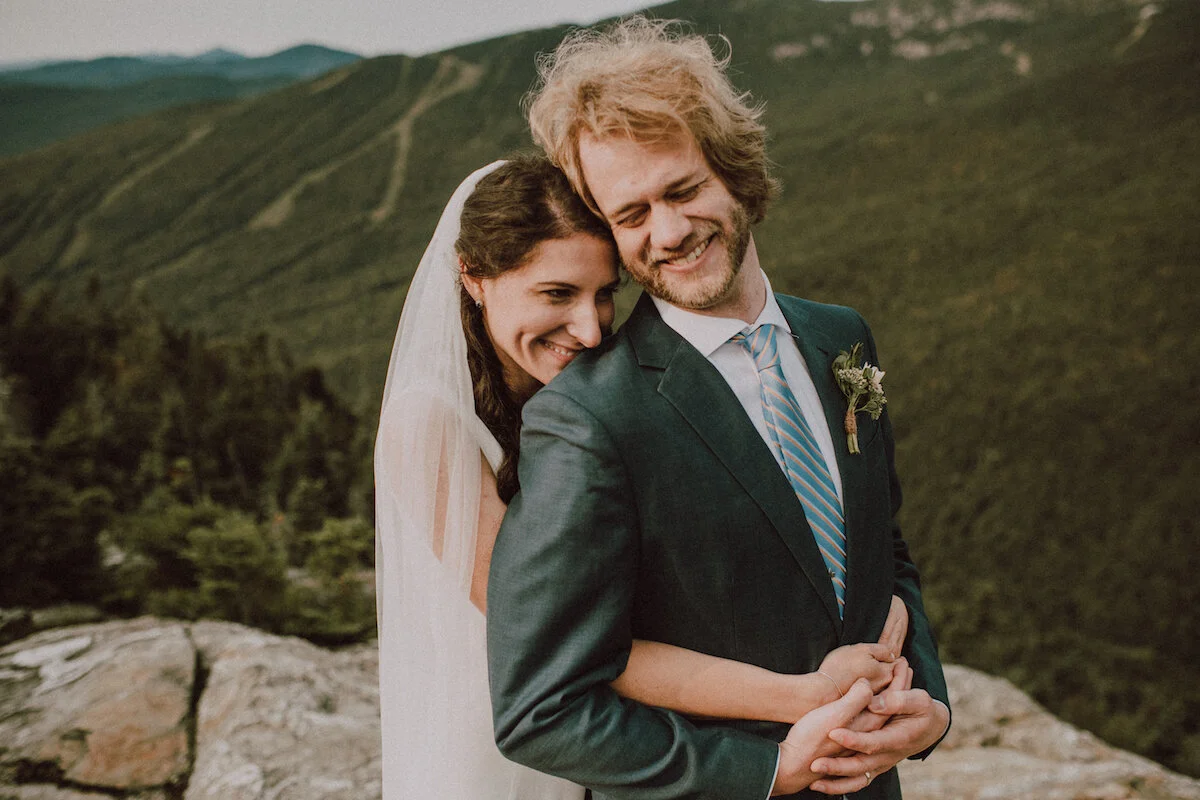 Bride hugs groom from behind on mountain