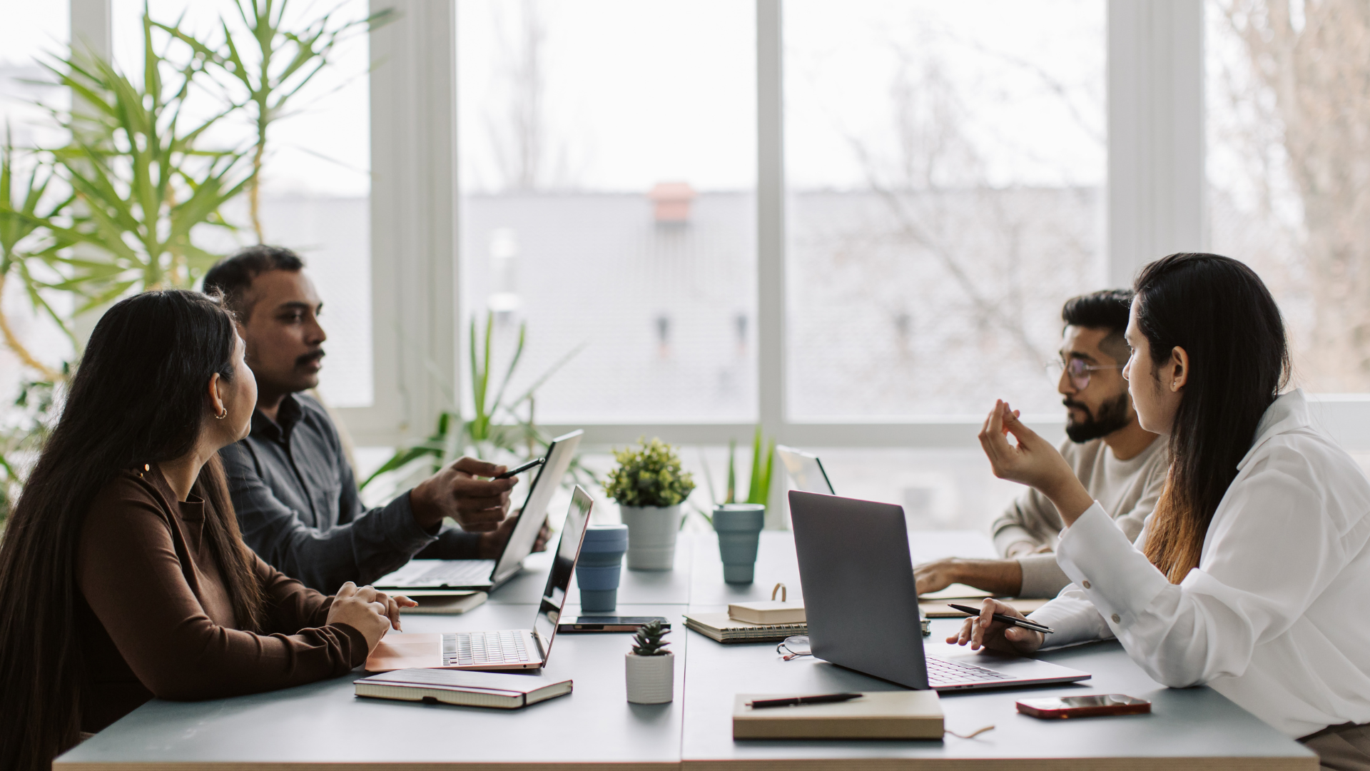 Four colleagues sitting across from each other in conversation at a table in a bright workplace.