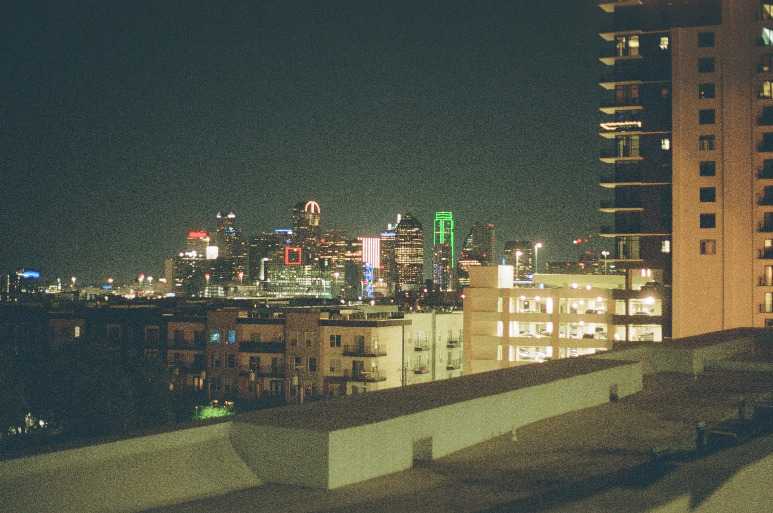 Nighttime city skyline with illuminated skyscrapers viewed from a balcony with residential buildings in the foreground. Photo of Dallas, Texas on 35mm Film. 
