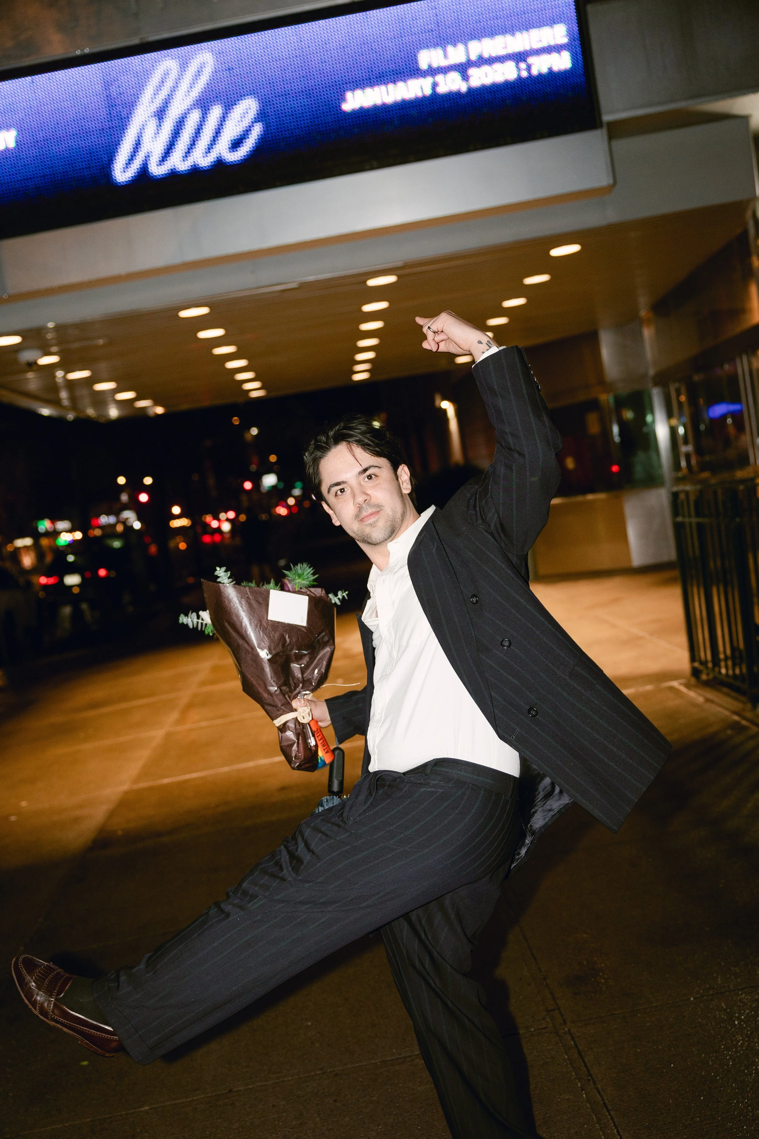 New York City Movie Premiere. New York Photographer. A man wearing a black pinstripe suit and white shirt, holding a bouquet of flowers, striking a playful pose in front of a theater marquee at night.