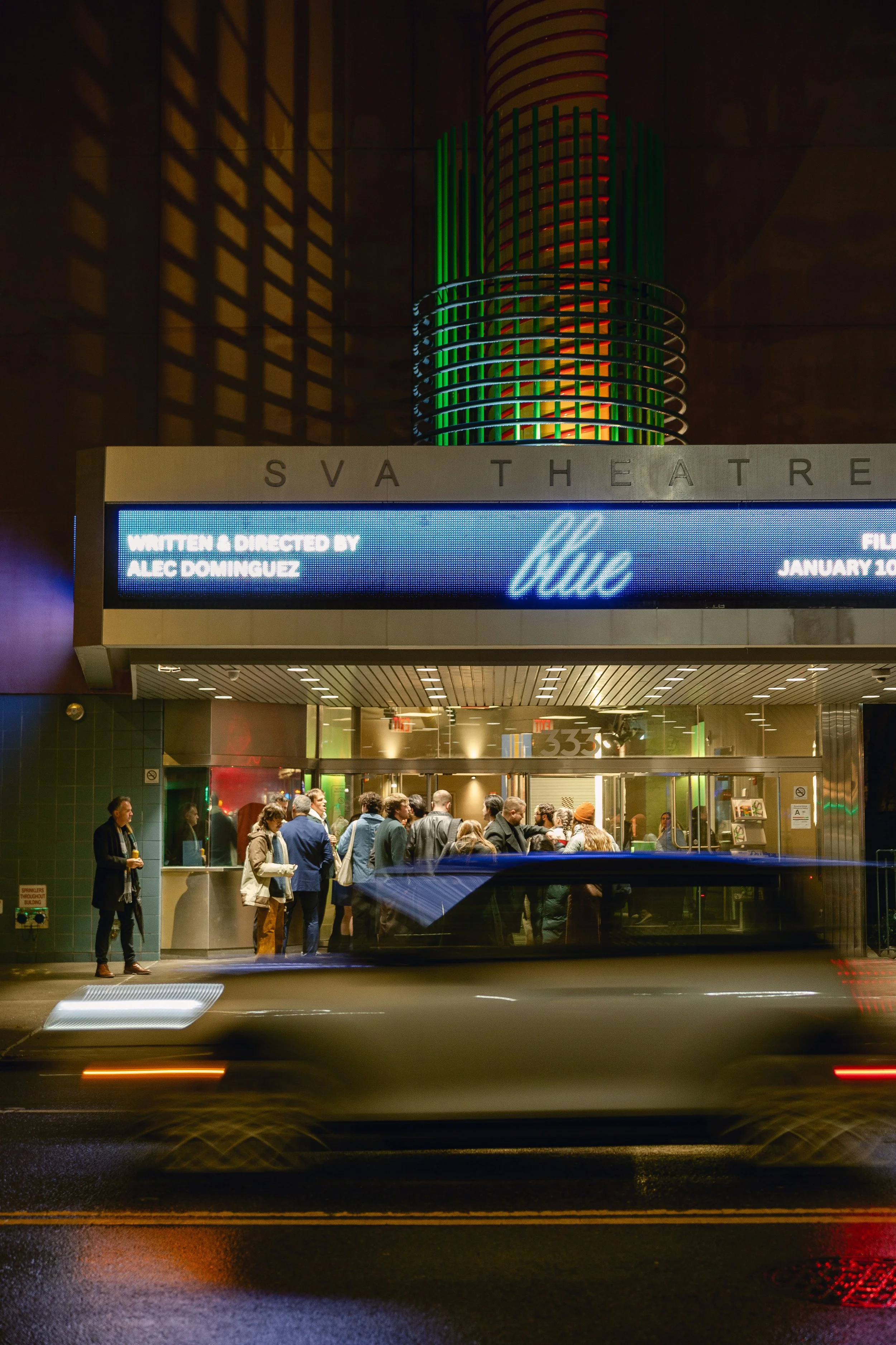 New York City Movie Premiere. People line up outside the SVA Theatre at night, with a digital marquee displaying the film title 'Blue' and the director's name, Alec Dominguez. A moving car passes in front of the scene, creating light streaks.