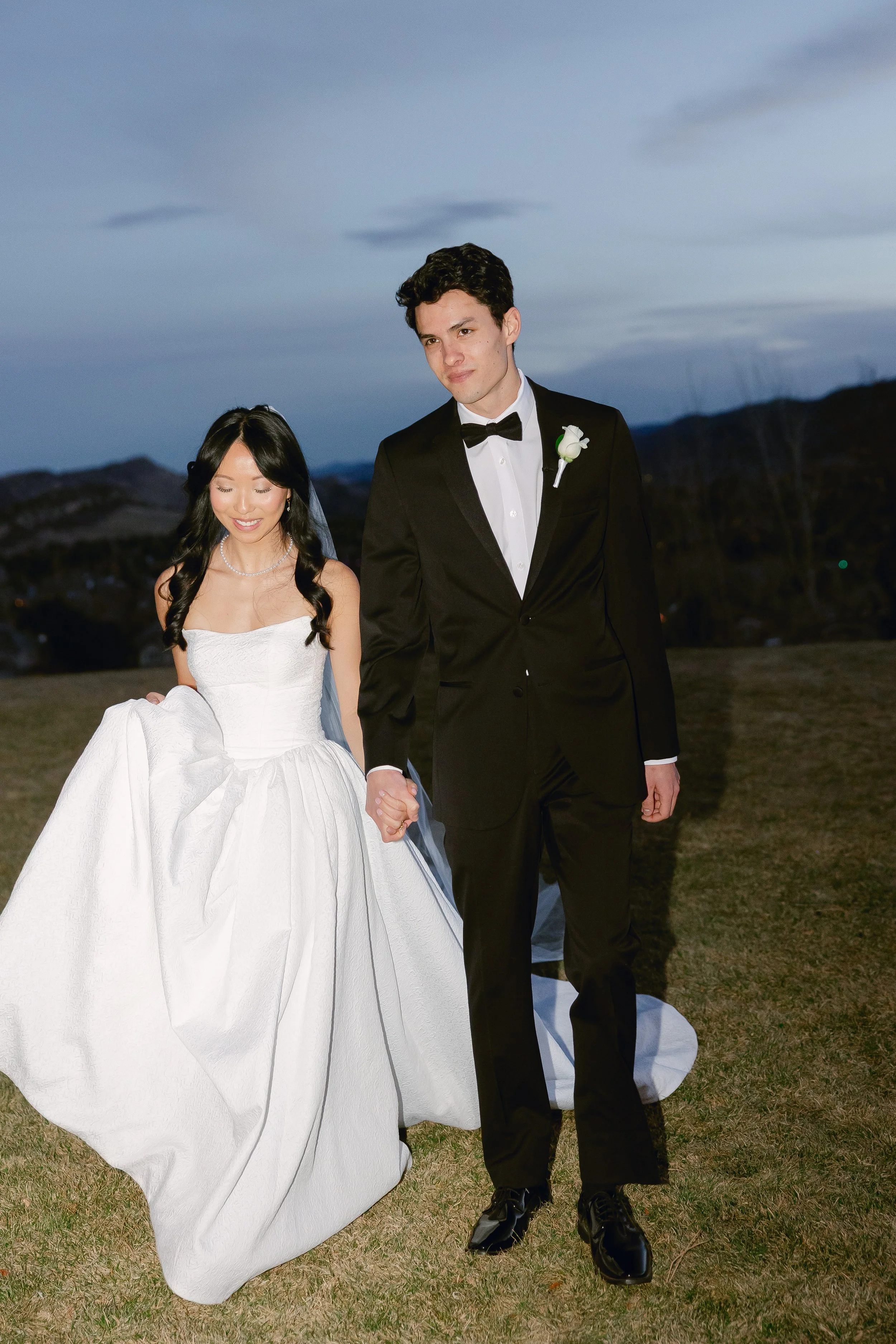 A newlywed couple walking hand in hand outdoors during dusk, with the woman in a white wedding gown and the man in a black tuxedo.
